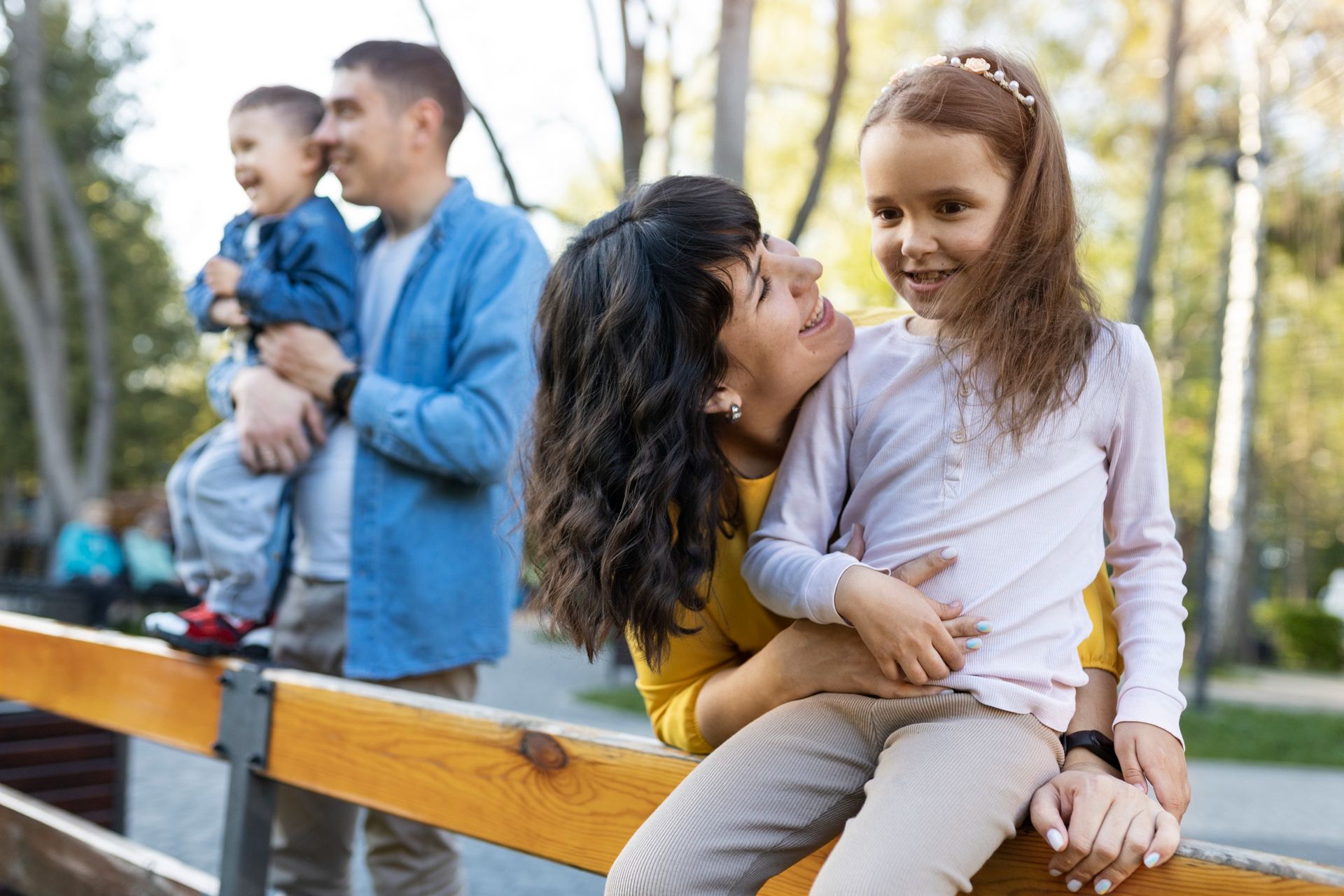 Two adults and two children resting on a park bench, smiling outdoors in sunlight.