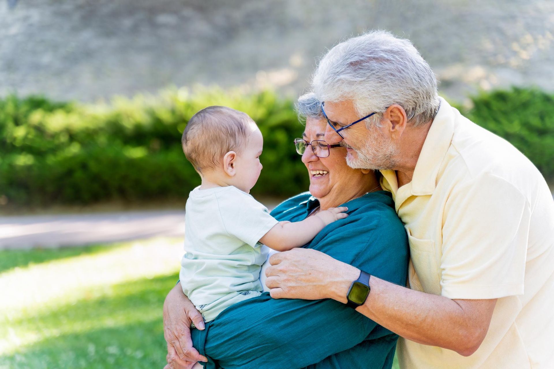 Older adult embracing a baby outdoors in a sunny park, both smiling beside green grass.