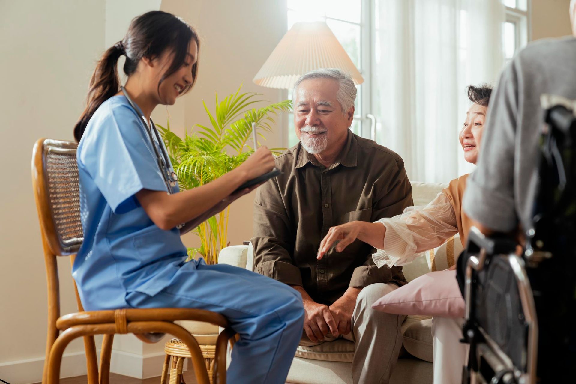 Caregiver in blue scrubs chatting with an elderly man and seated woman in a bright living room