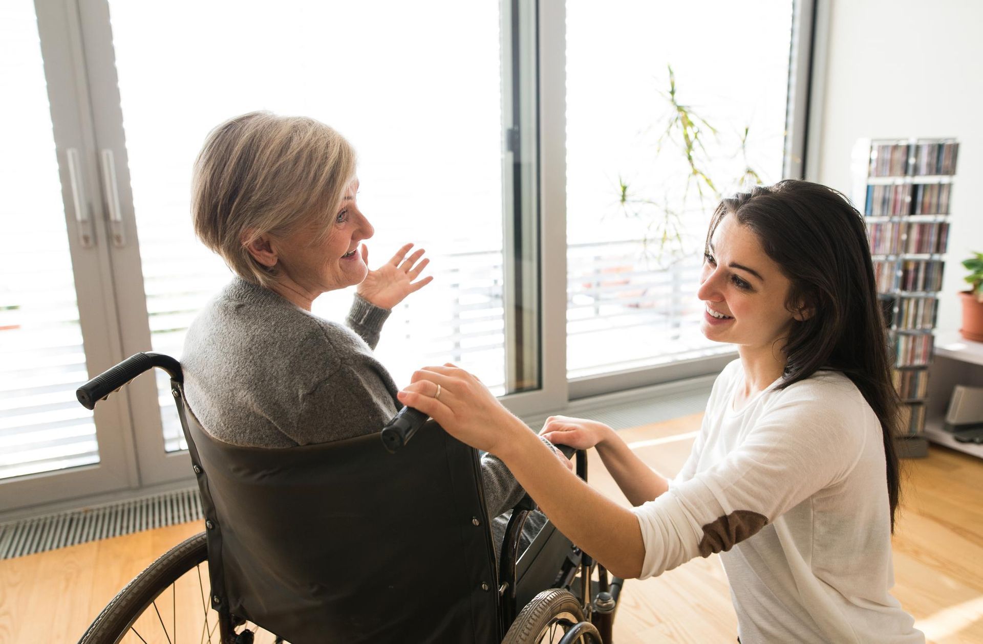 Two women chatting indoors, one seated in a wheelchair near a bright window