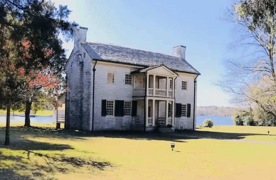 White historic house with black shutters on a sunny lawn by water, framed by trees