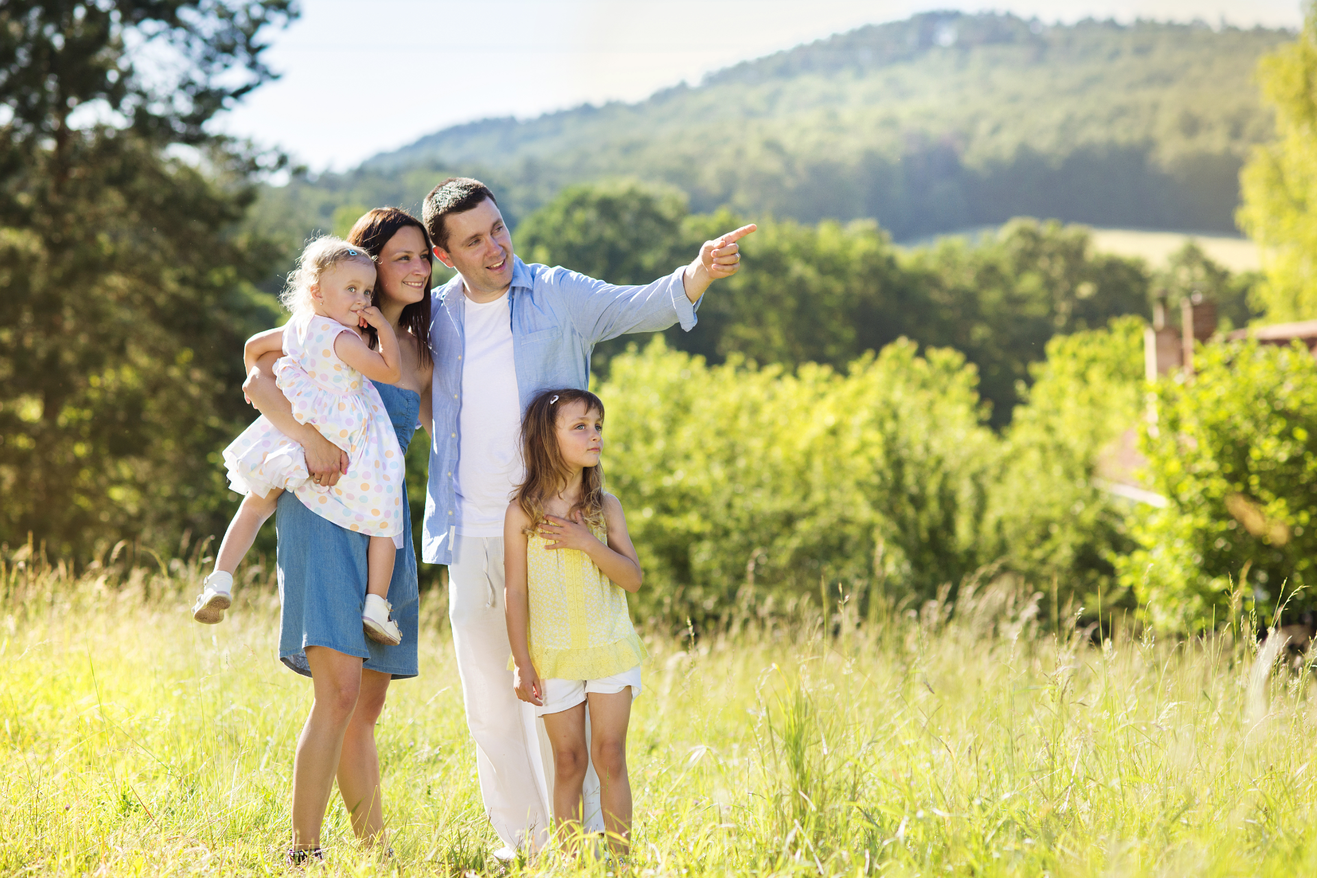 Family of four standing in a sunny meadow, with a man pointing toward distant hills.