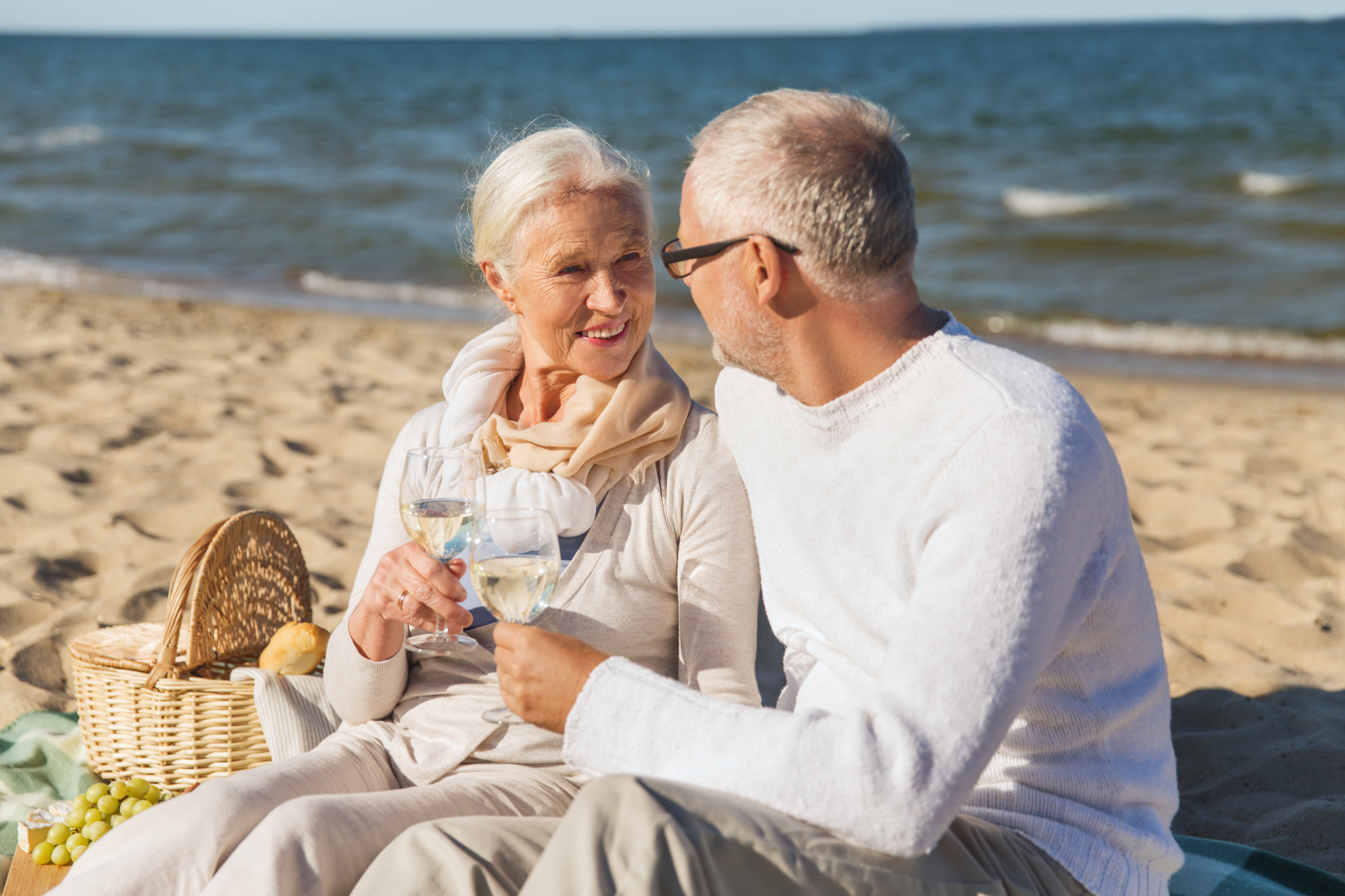 Two people sitting on a beach blanket, smiling and talking beside a picnic basket by the sea