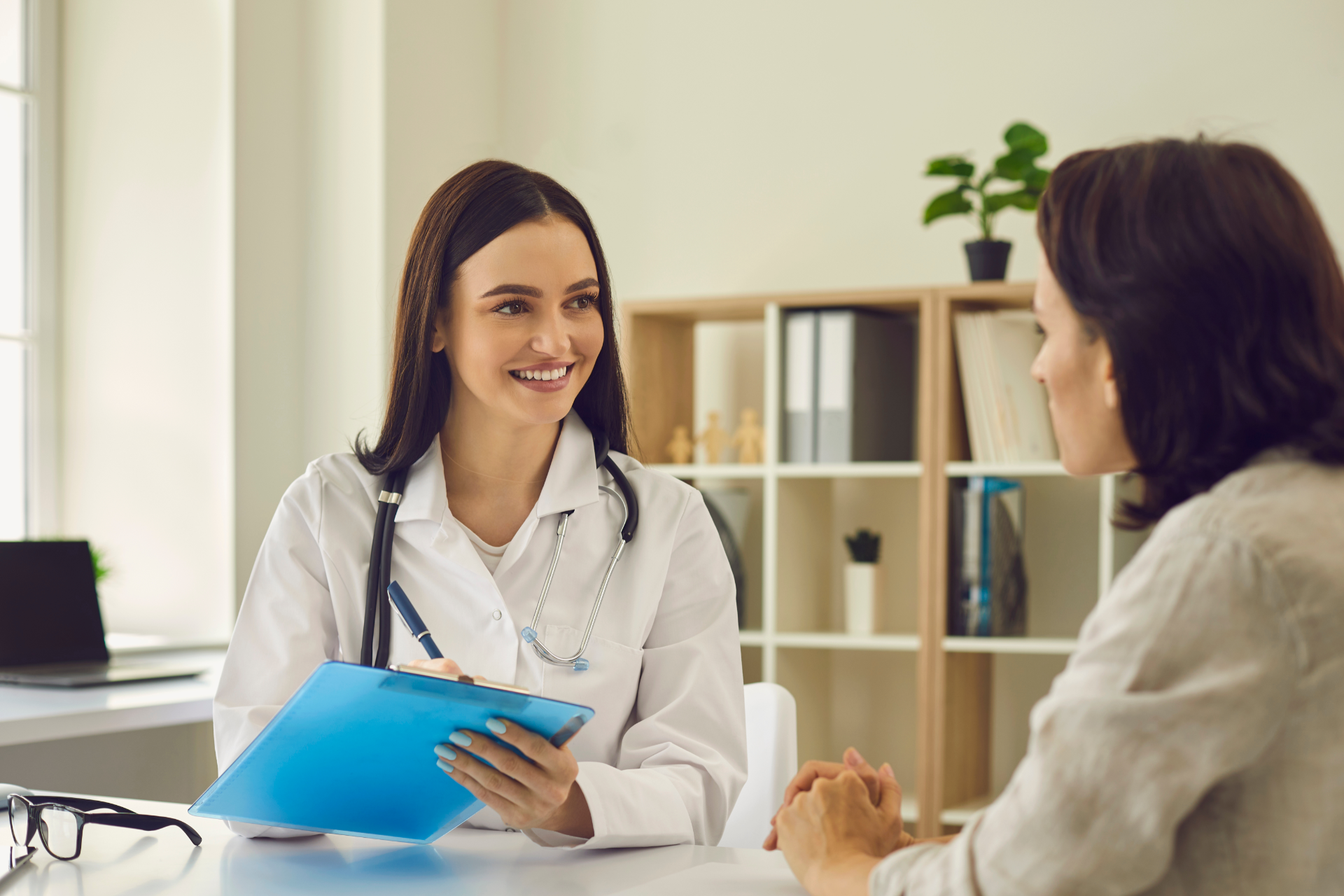 Doctor smiling with a patient in a bright office, holding a blue clipboard during a consultation