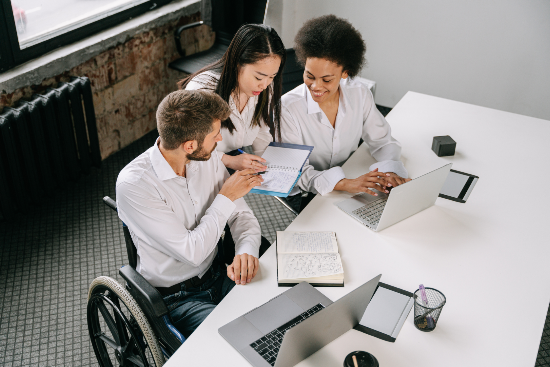 Three coworkers in white shirts discussing documents at a bright office table with laptops and a wheelchair user