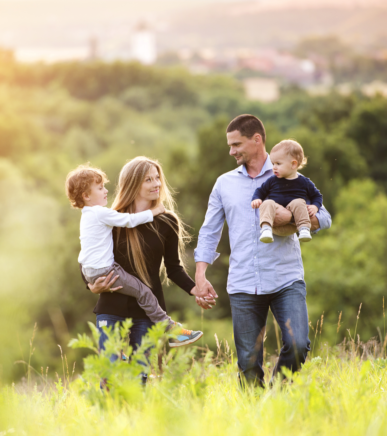 Family walking hand in hand through a sunlit grassy field, with two children and two adults smiling.