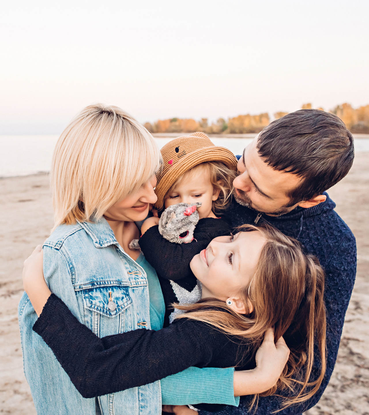 Family hugging by a lakeshore, smiling and cuddling close in soft autumn light