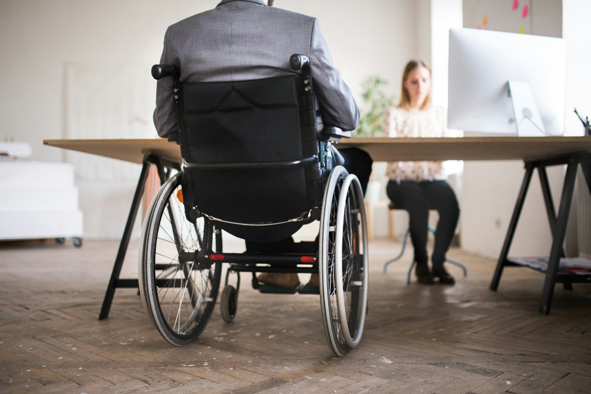 Person in a wheelchair facing a desk in an office, with a woman seated behind it.