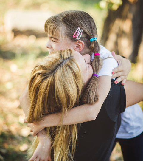 Woman hugs smiling child outdoors in a sunlit park, with colorful hair clips visible.