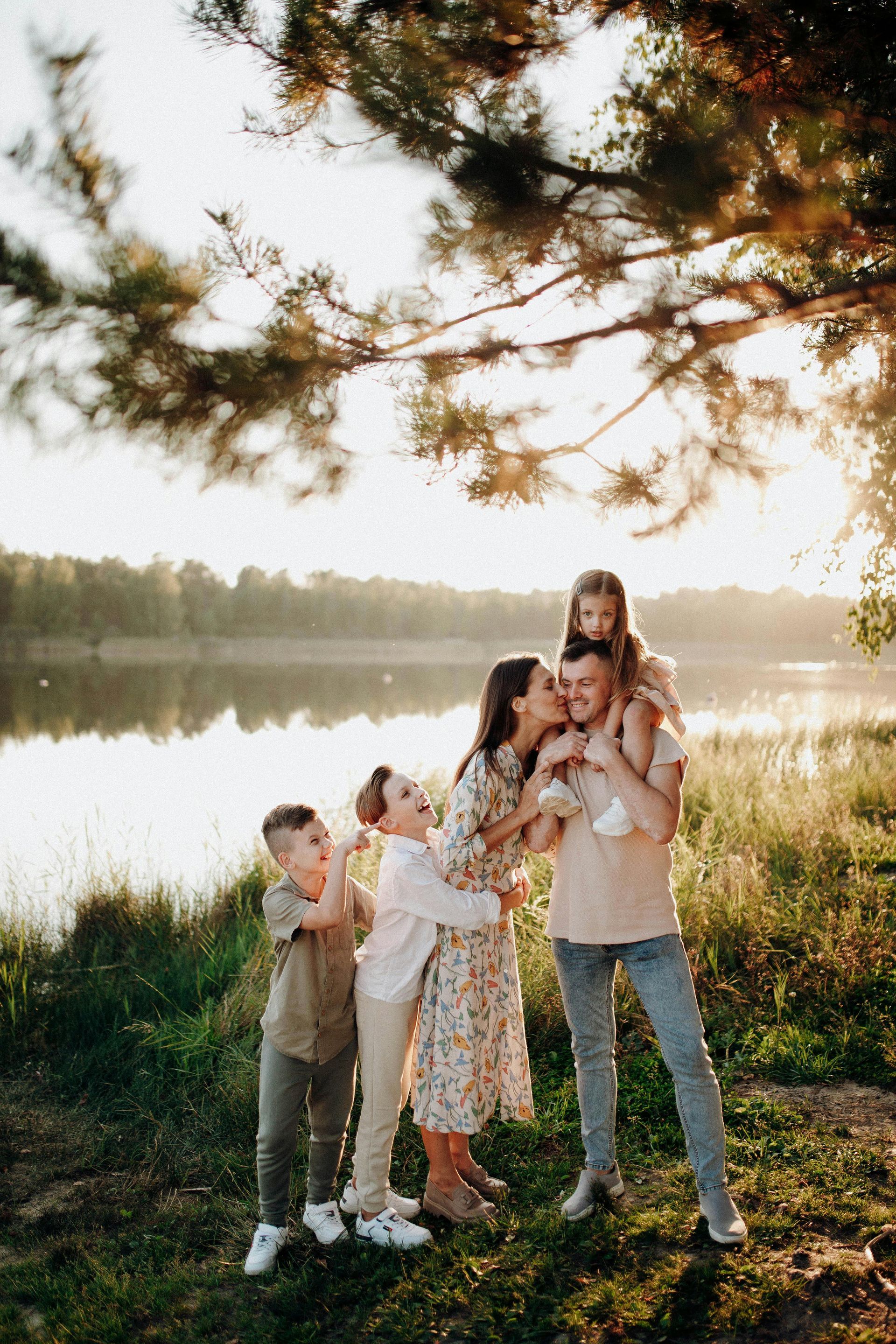 Family standing by a lakeside at sunset, with two children and two adults beneath overhanging branches.