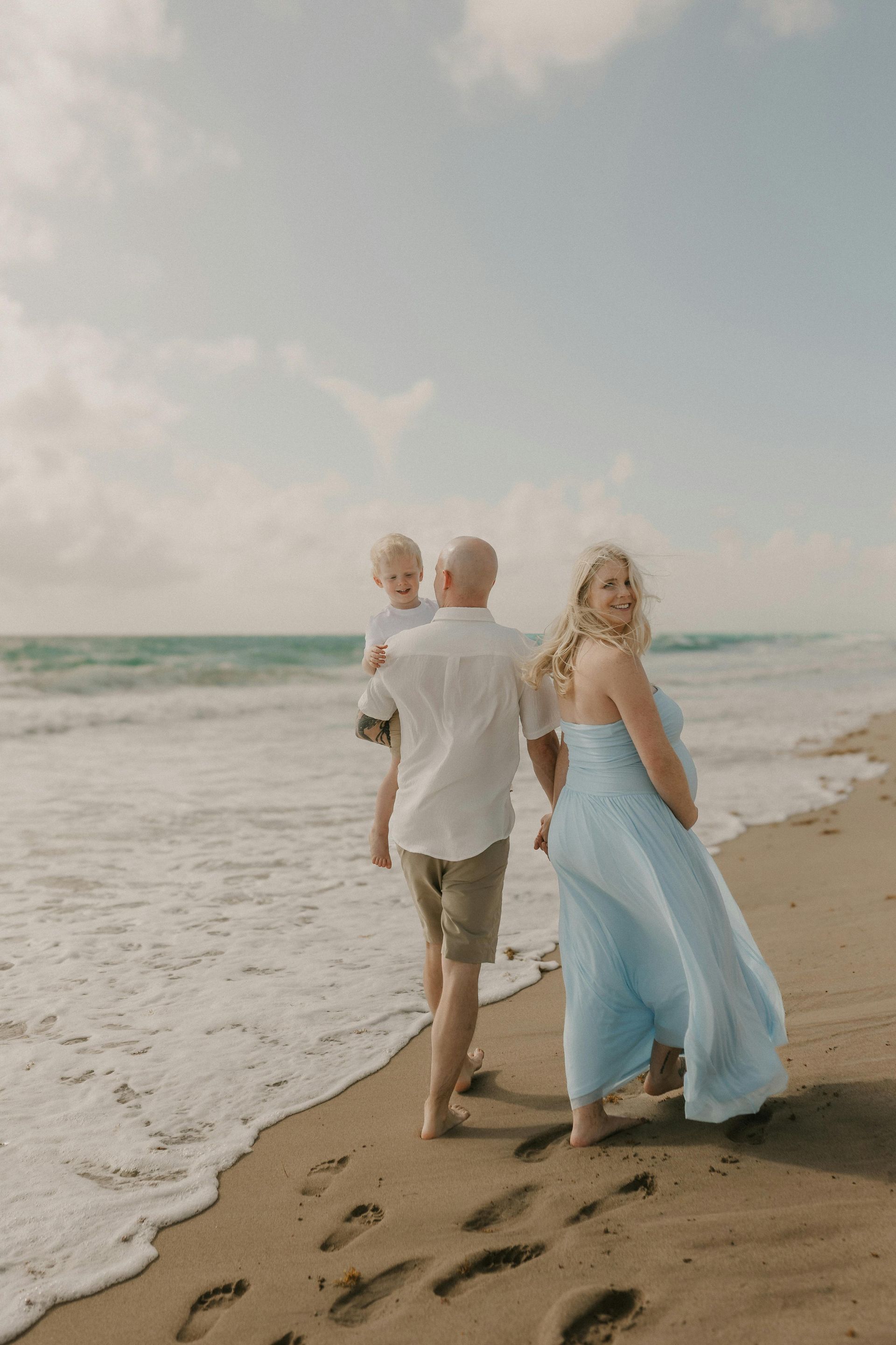 Two people walking along a sandy beach beside foamy waves, one in a white shirt and one in a blue dress.