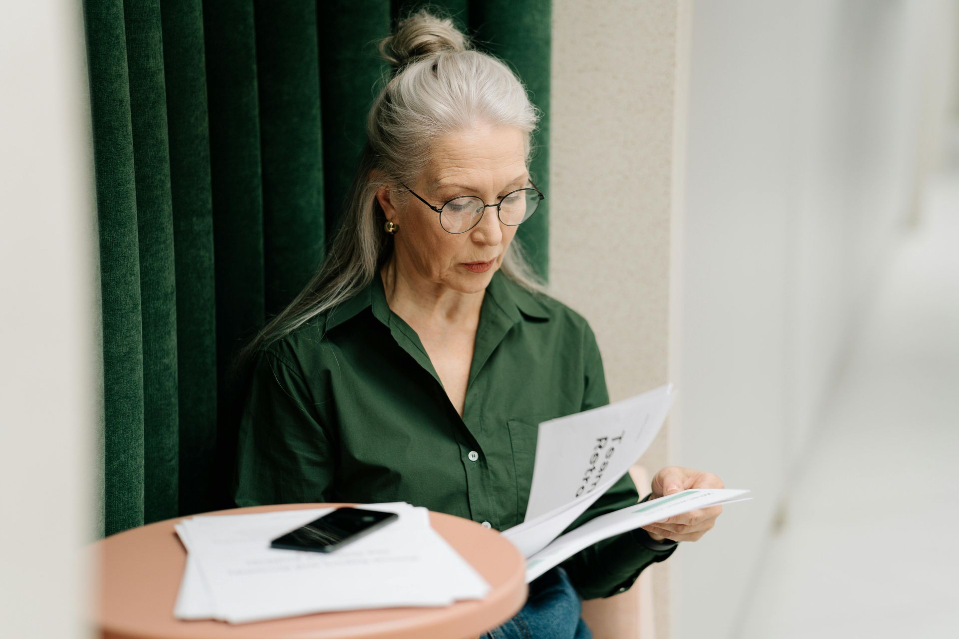Woman in a green shirt reading papers beside a table with a black phone in a softly lit room