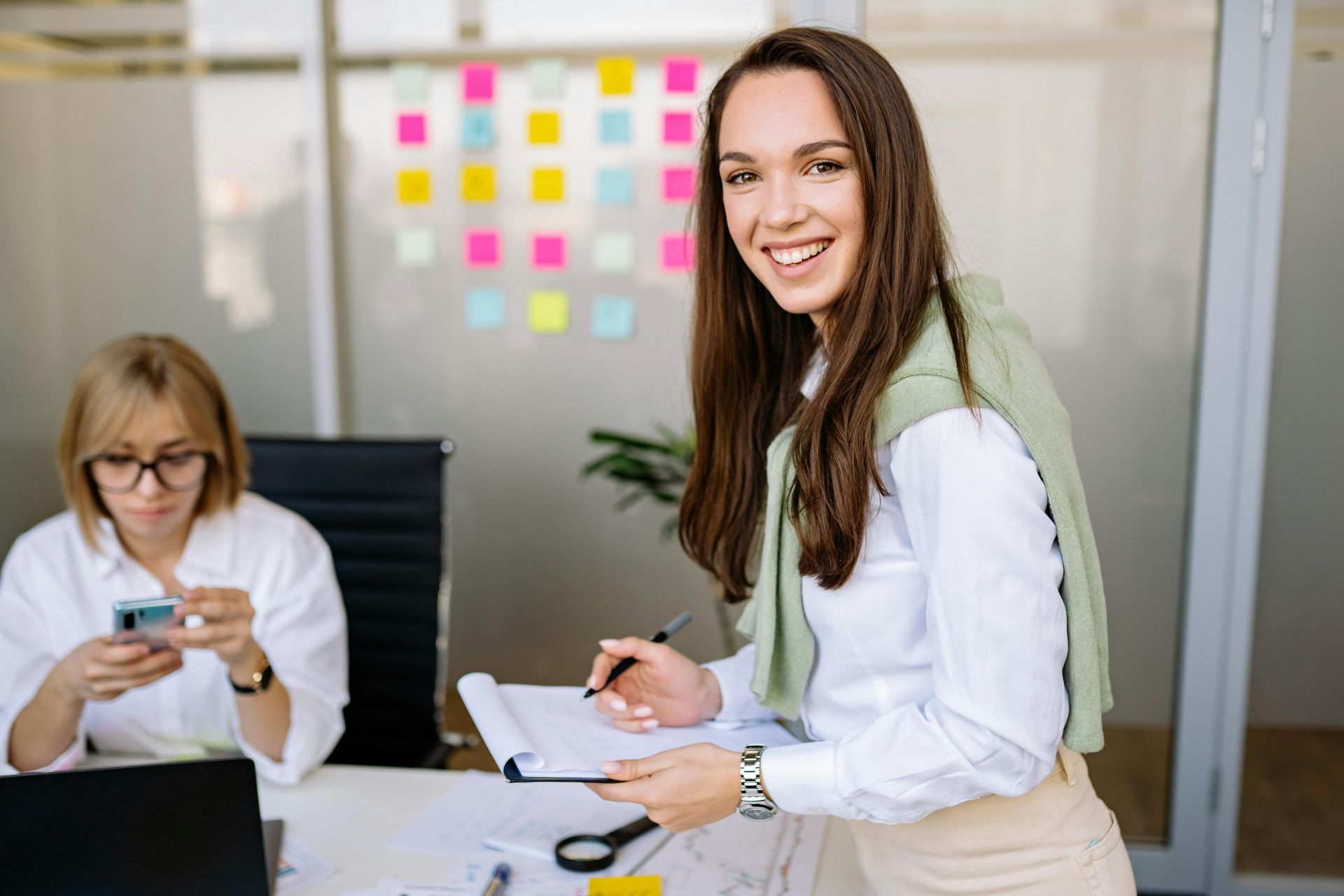 Two women in an office, one smiling at camera while holding papers, another looking at her phone near a laptop.