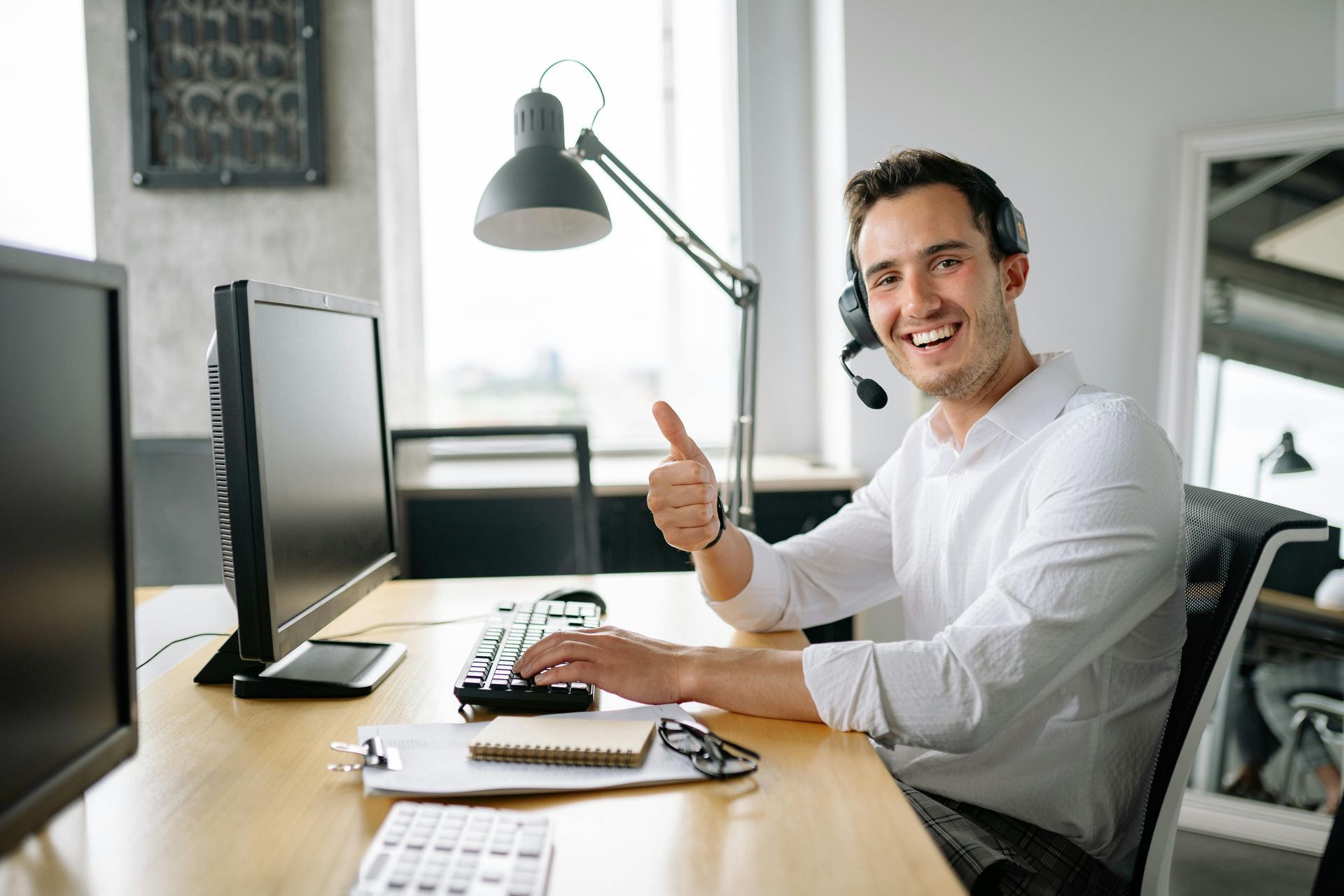 Smiling man in a white shirt at a desk, gesturing in a bright office with a computer and headset