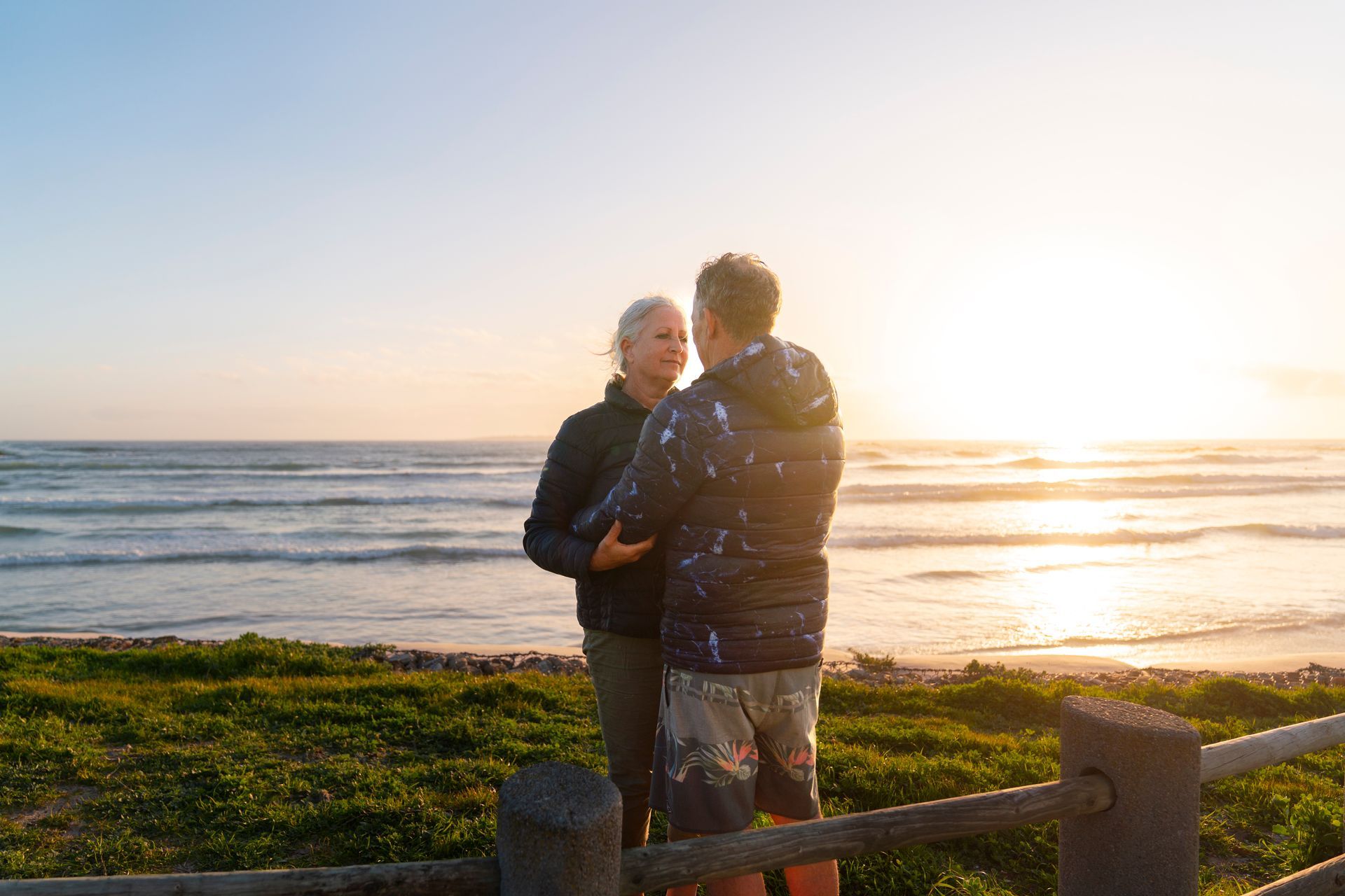 Two people stand on a beach at sunset, facing each other by the shoreline.