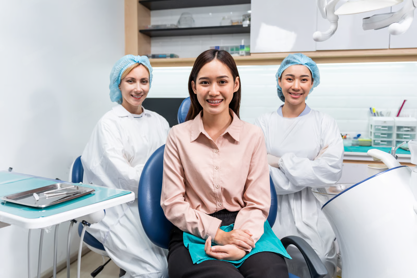 Dentist’s office with a smiling patient seated between two clinicians in white coats and blue caps