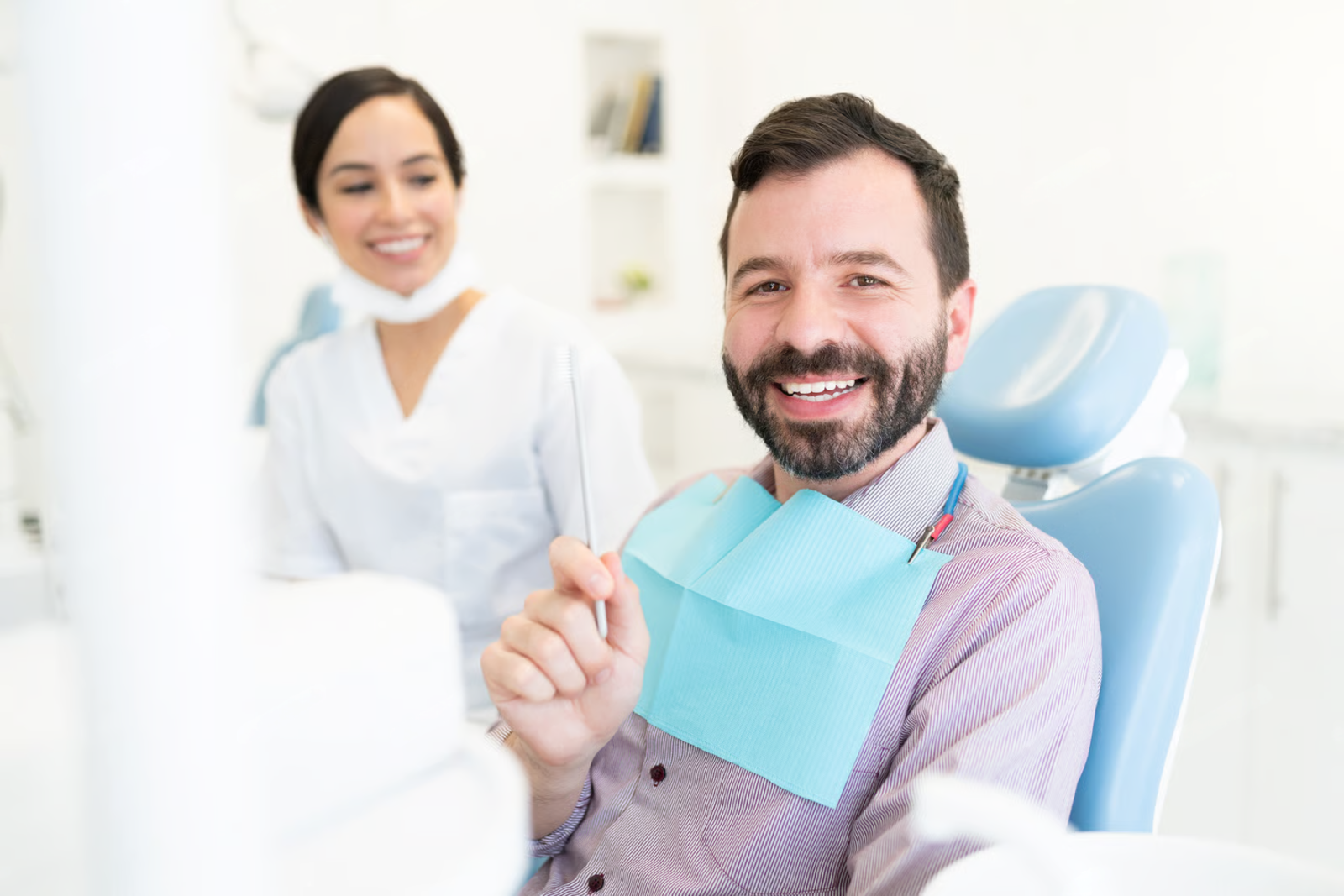 Smiling patient in a dental chair holding a blue toothbrush, with a dentist in the background