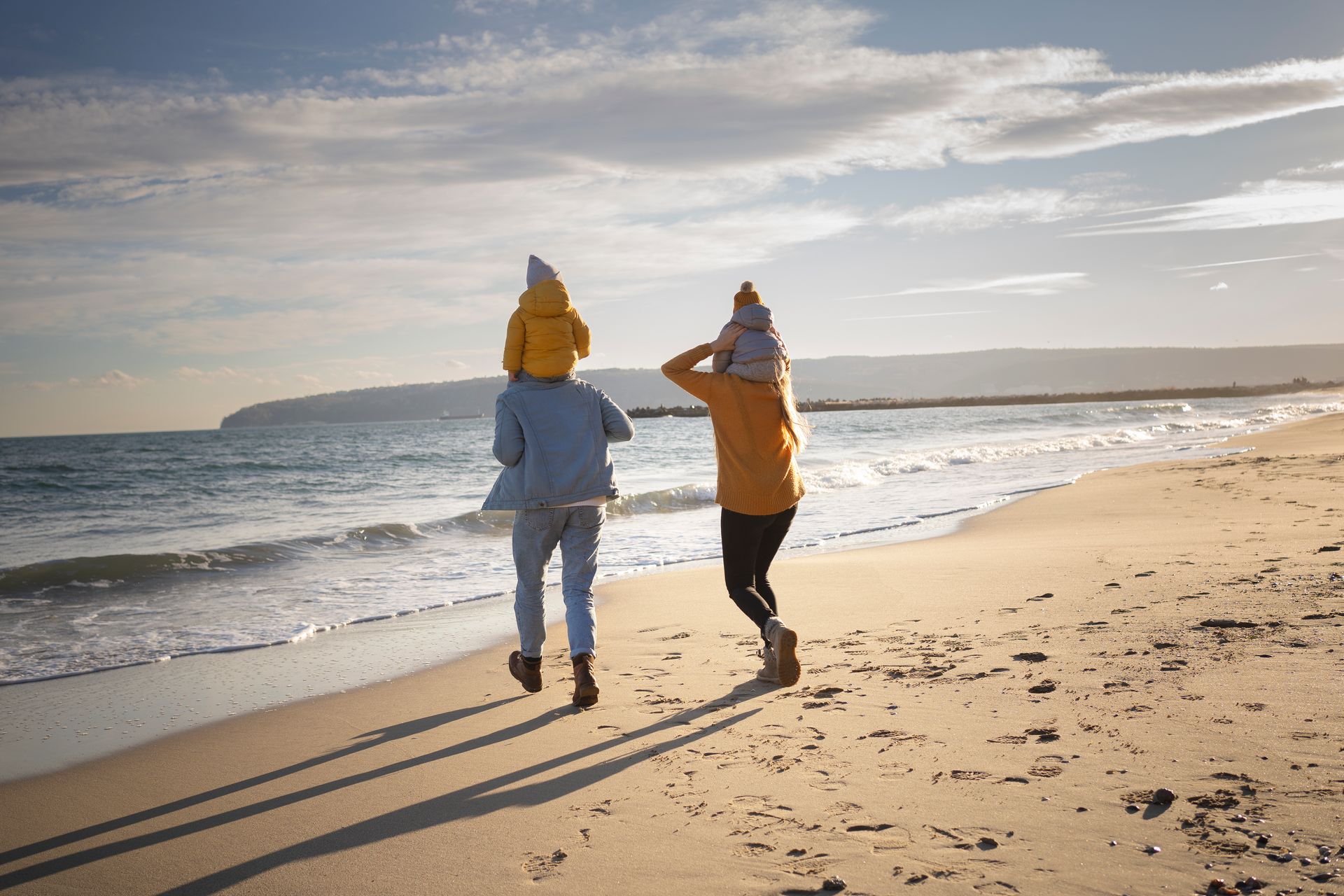 Two people walking along a sunny beach, one in blue and one in orange, with waves and a distant headland.