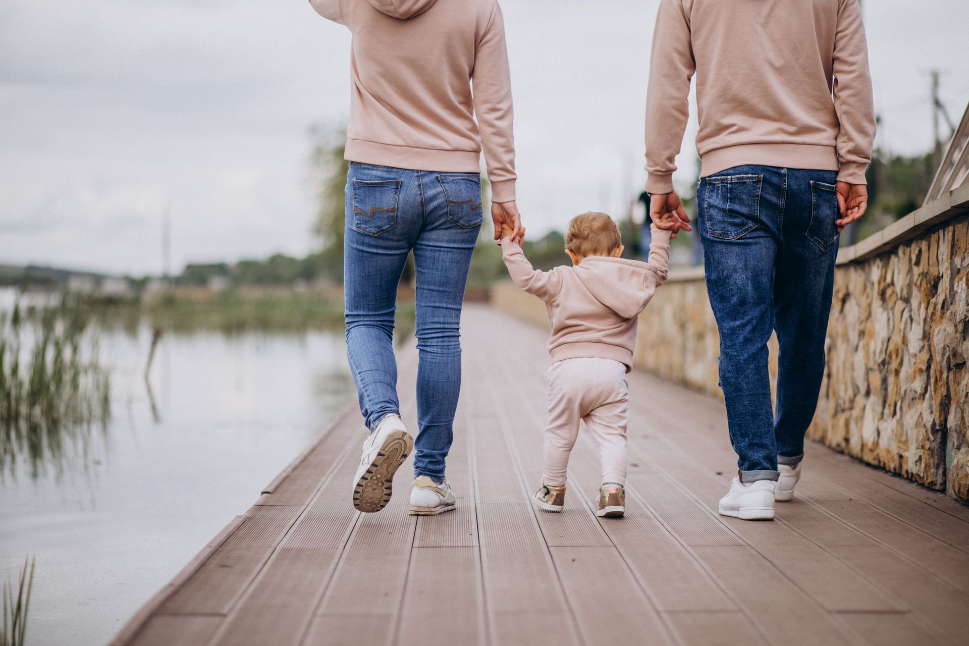 Two adults holding a toddler’s hands while walking on a wooden path beside the water