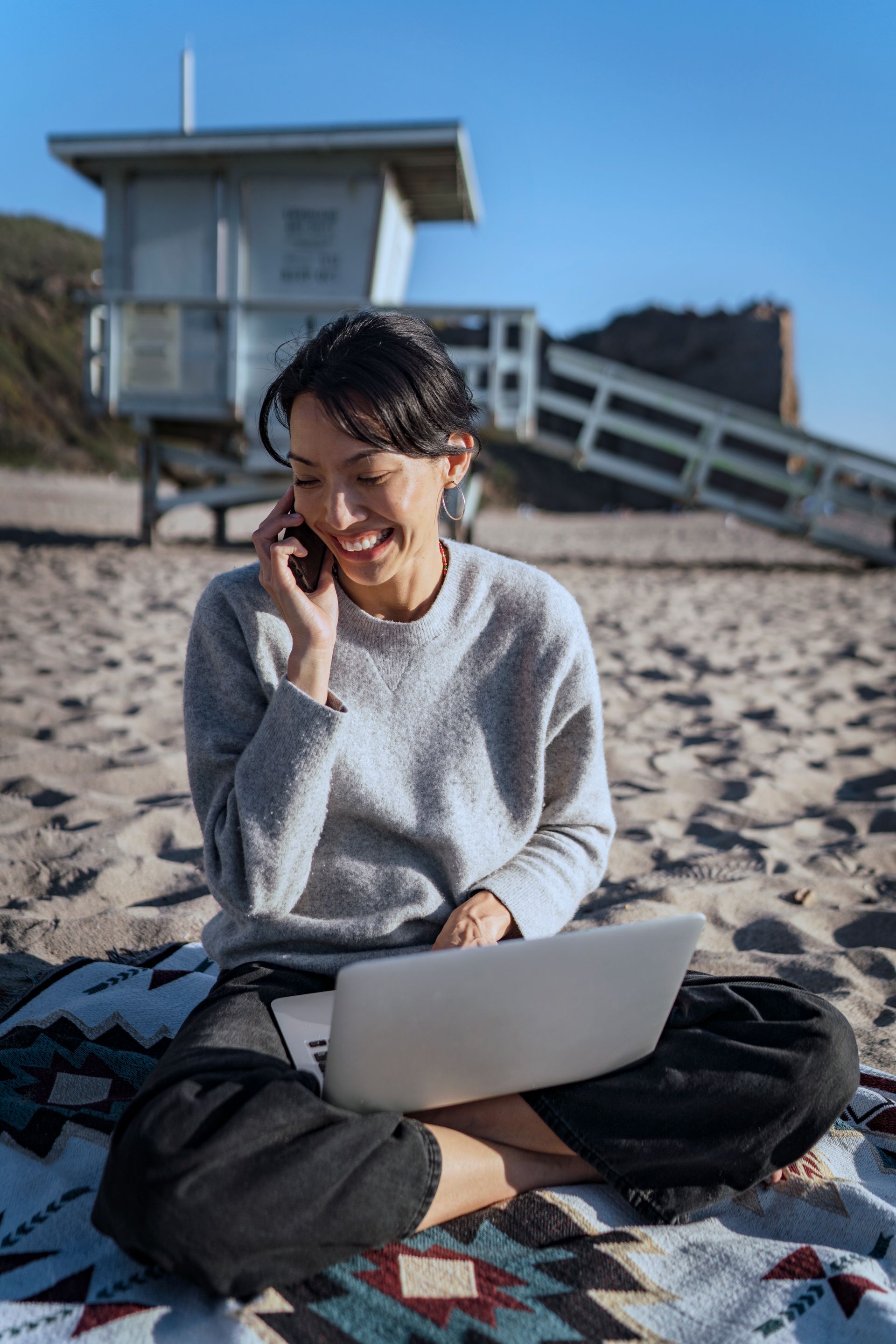 Woman smiling on a beach, using a laptop and phone beside a lifeguard tower