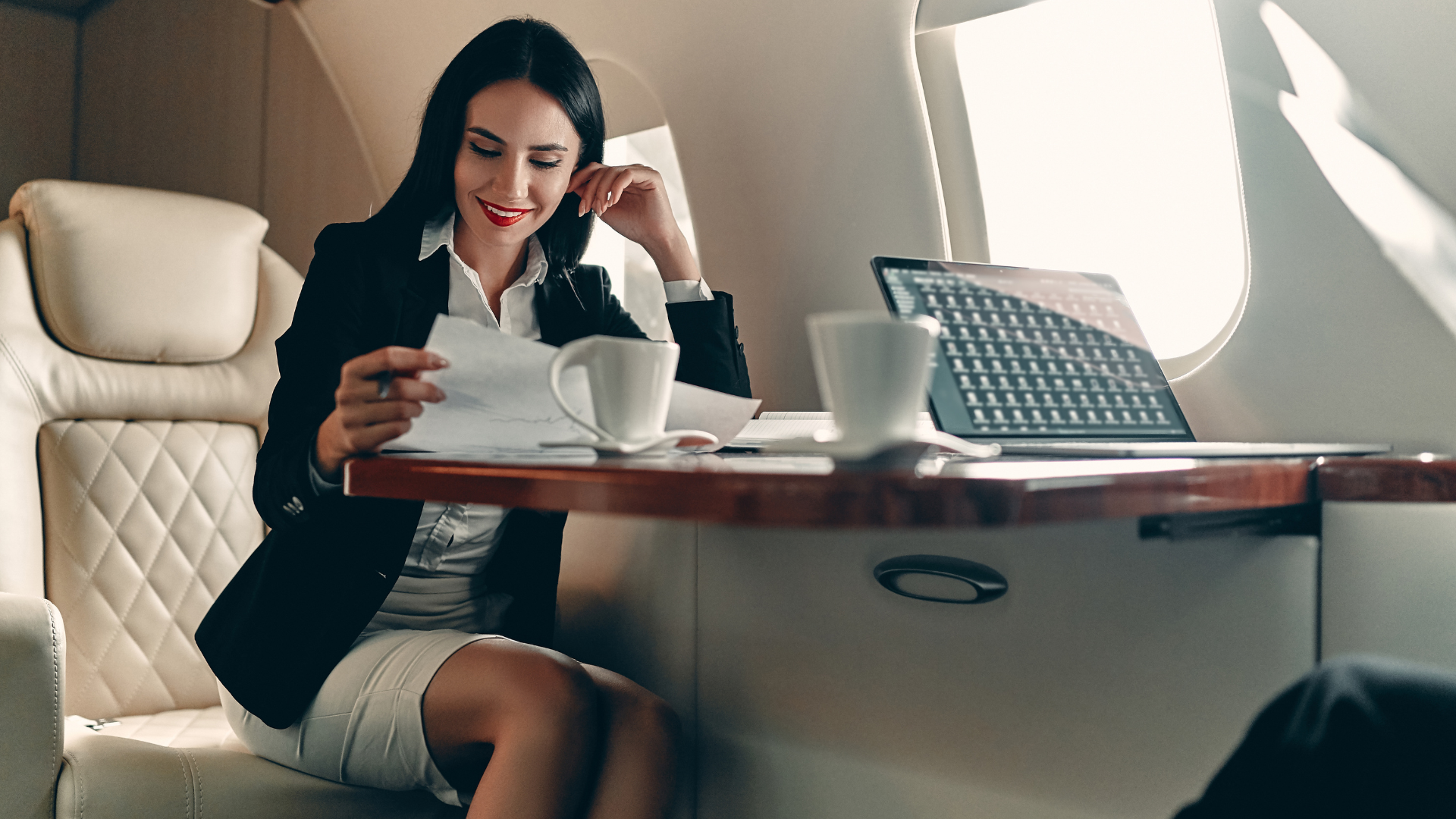 Woman smiling at a laptop on an airplane, with coffee cups on the tray table.