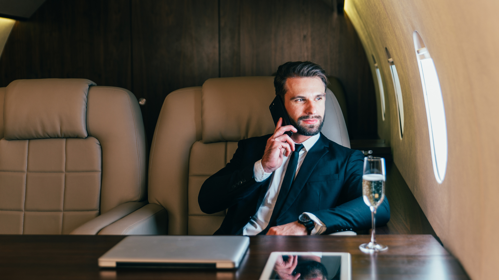 Man seated in a private jet cabin, speaking on a phone beside a table and champagne glass
