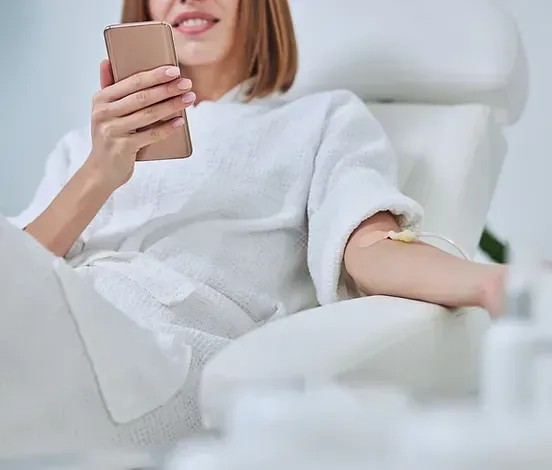 Woman in white robe receives IV drip while using a phone, sitting in a chair.