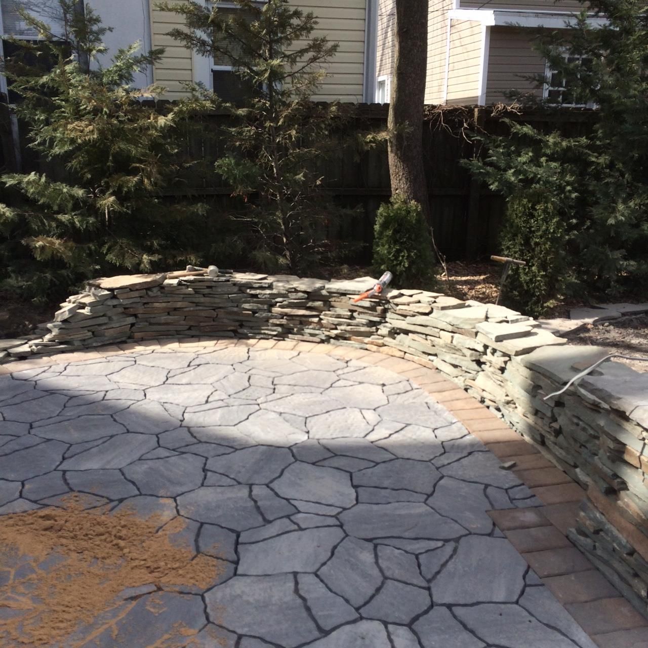 Stone-paved backyard patio in Millstone NJ with curved retaining wall, trees, and house in the background