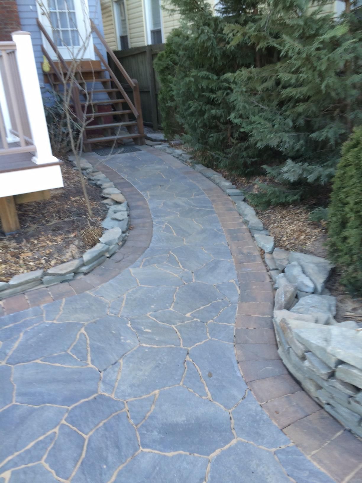 Stone walkway in Colts Neck curving beside a house, bordered by brick and rocks, leading toward stairs and shrubs