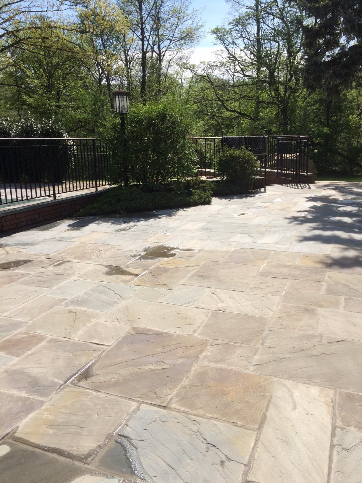 Stone patio in Middletown NJ with black railings, green trees, and a small garden under bright sunlight