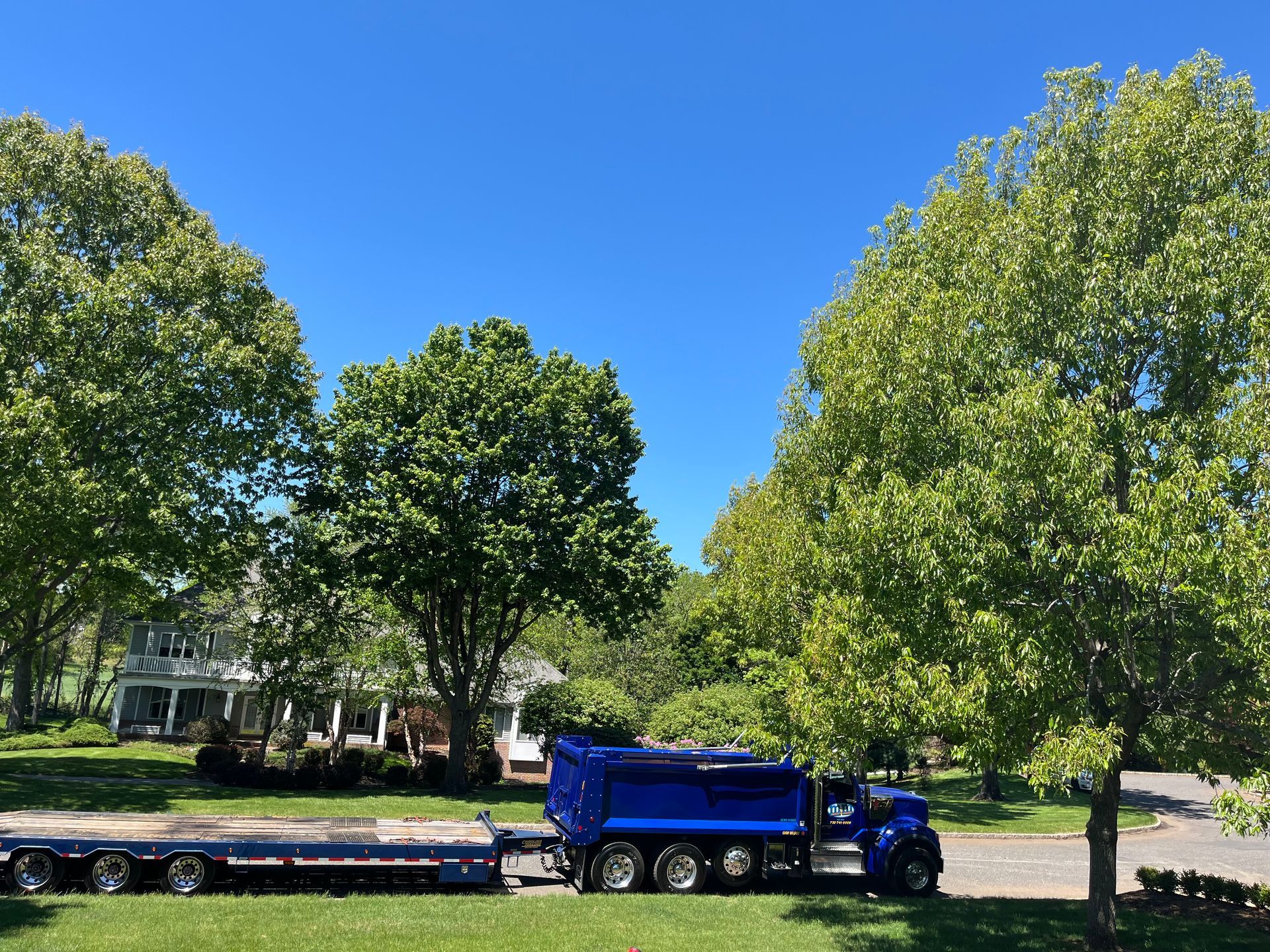Driveway Paving Contractor Colts Neck. NJ. Blue dump truck with flatbed trailer parked on a tree-lined street under a clear blue sky