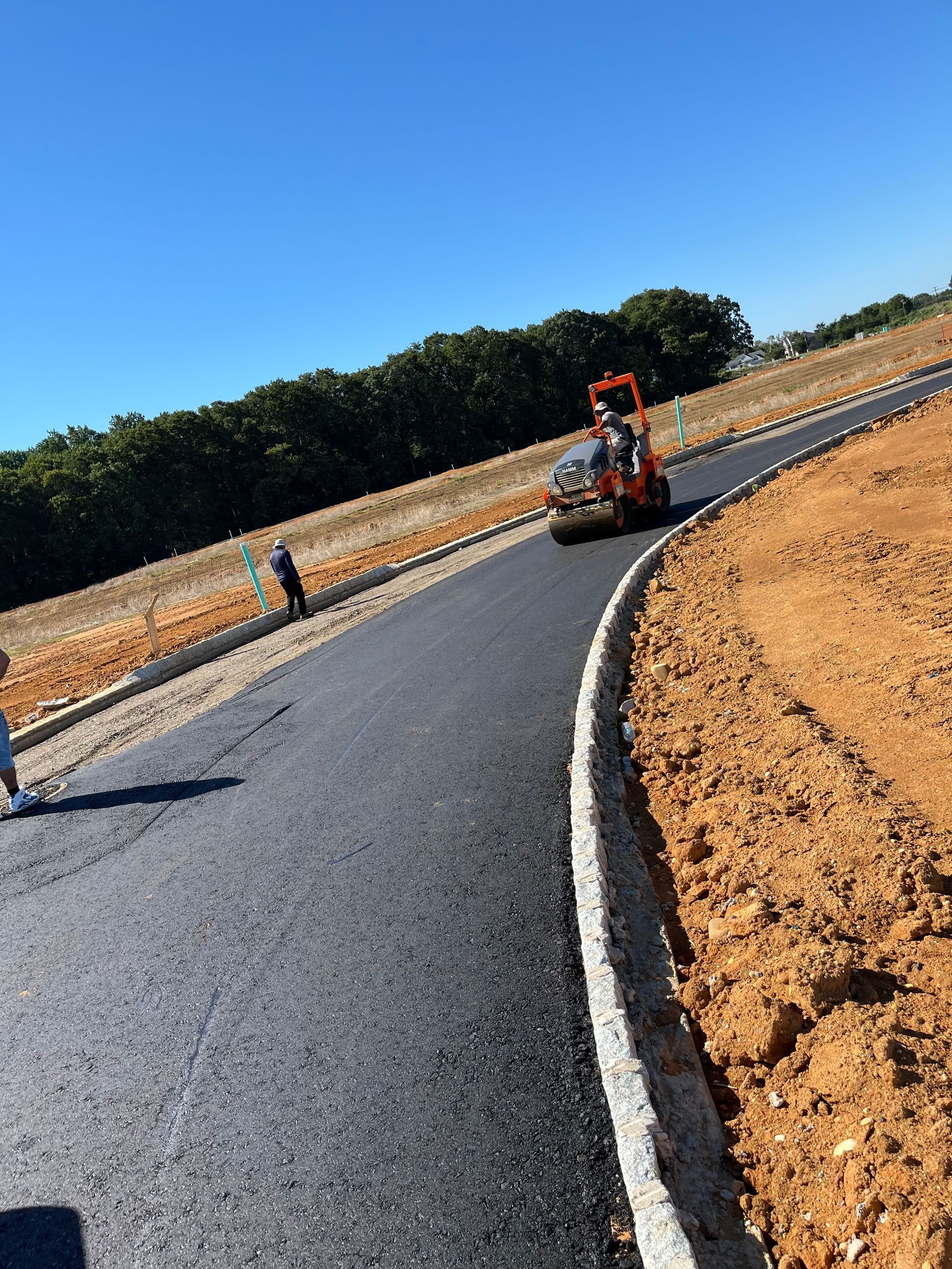 Millstone Paving services. Curving fresh asphalt road under construction with a roller on the bend and a worker nearby under a blue sky