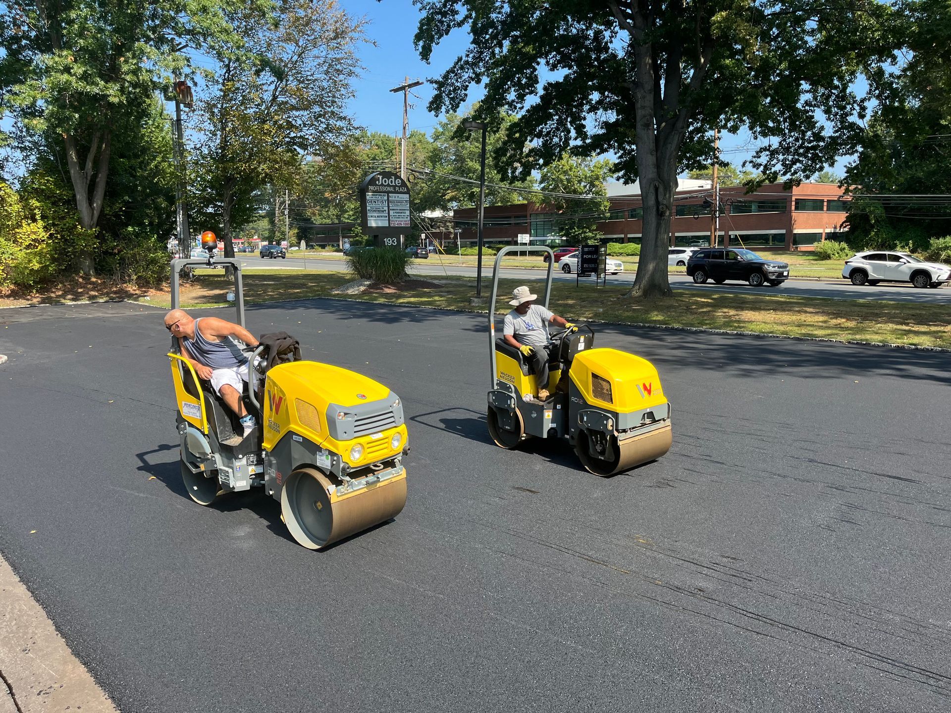 Best Paving Company in Rumson NJ. Two workers ride yellow road rollers on a paved street lined with trees and parked cars.