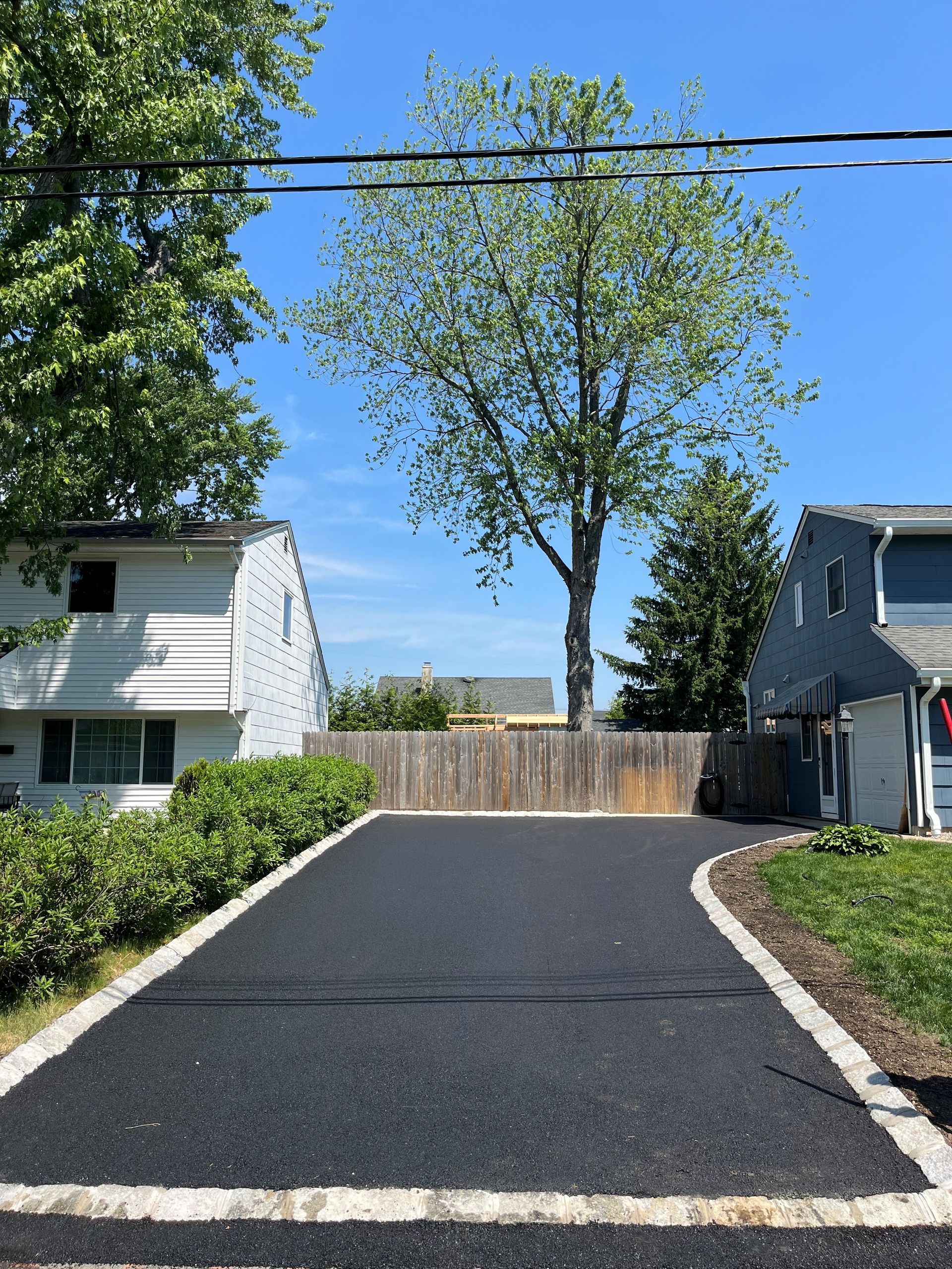 Best Blacktop Paving in Millstone NJ. Freshly paved driveway between two houses, leading to a wooden fence and a tall tree
