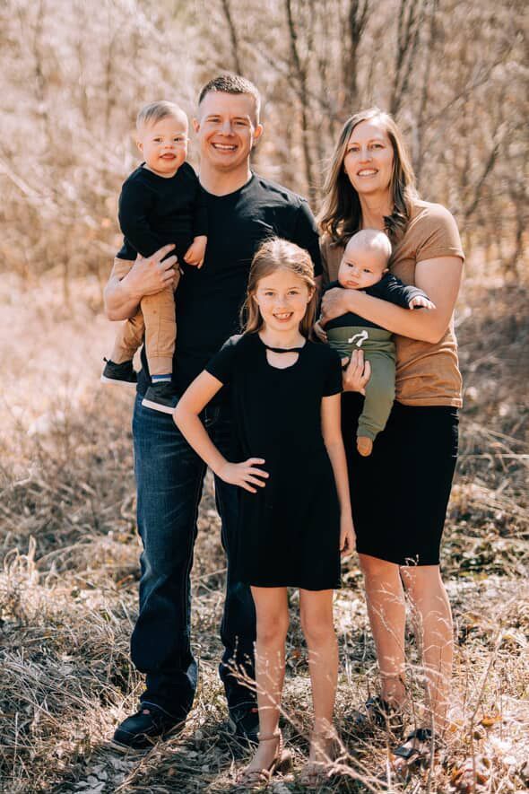 Family of five smiling outdoors. Man holds a child with Down syndrome, woman holds a baby, child stands in front.