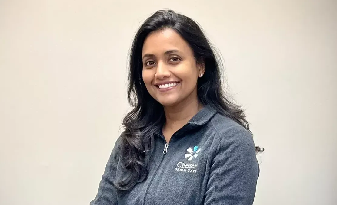 Woman with long dark hair, wearing a gray zip-up sweatshirt, smiles at the camera against a plain background.