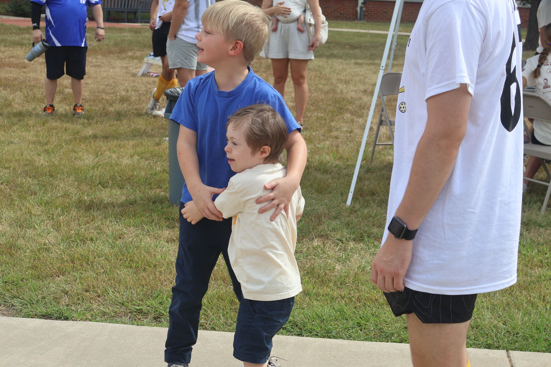 Boy in blue shirt hugs a toddler with Down syndrome; people watch on a grassy area, sunny day.