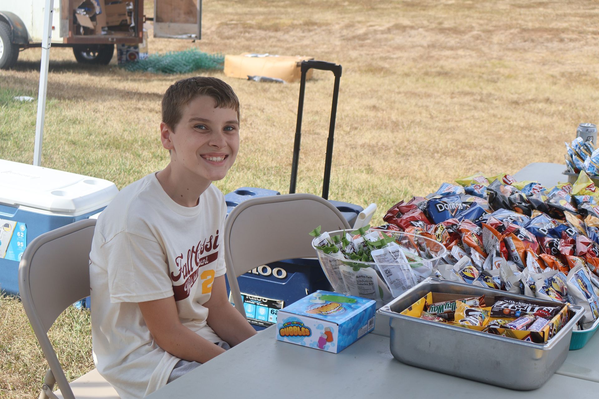 Boy smiling at a table selling snacks and toys outdoors.