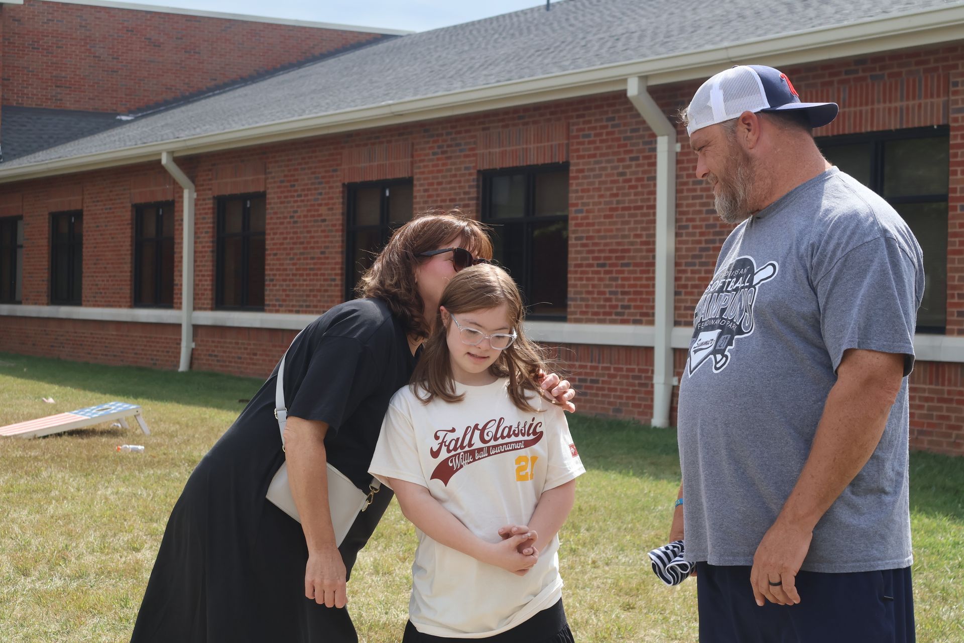 Woman hugs a girl with Down syndrome, man watches, outdoors near a brick building.