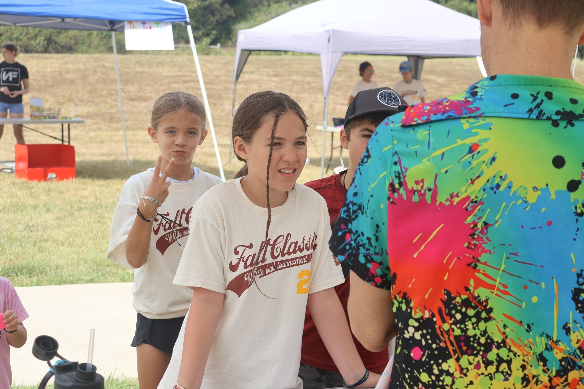 Children at an outdoor event. Two girls in matching shirts stand near a person in a colorful shirt.