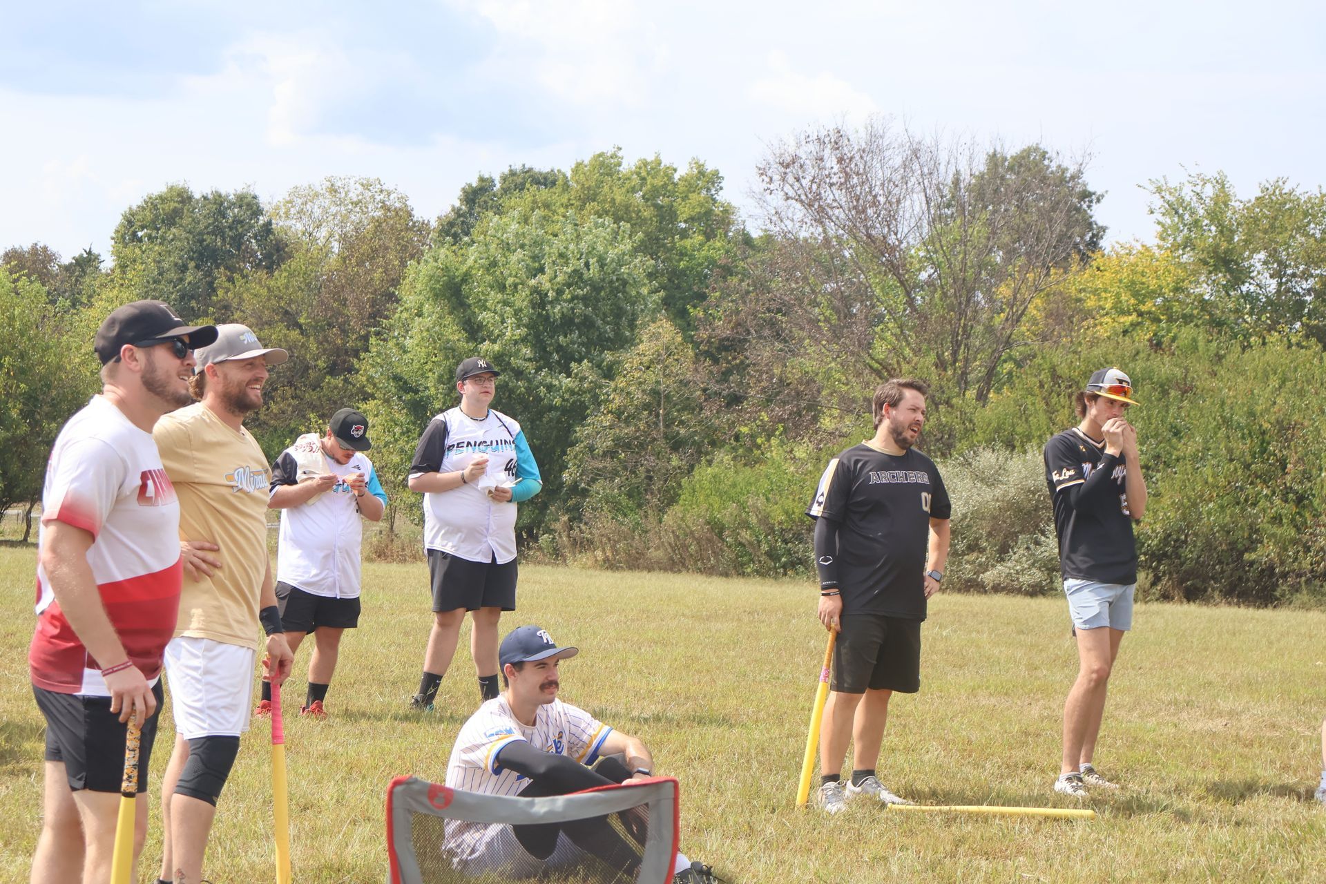 Group of people watching a game on a grassy field. Some are holding wiffleball bats, and a net is in foreground.