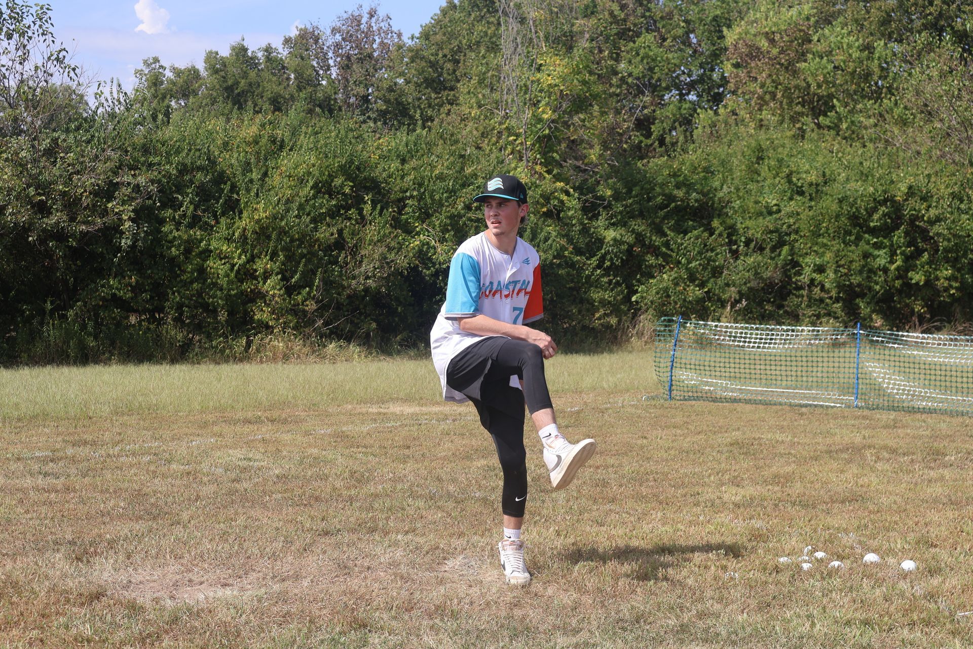Person in wiffleball uniform, mid-pitch on a grassy field with trees in background.