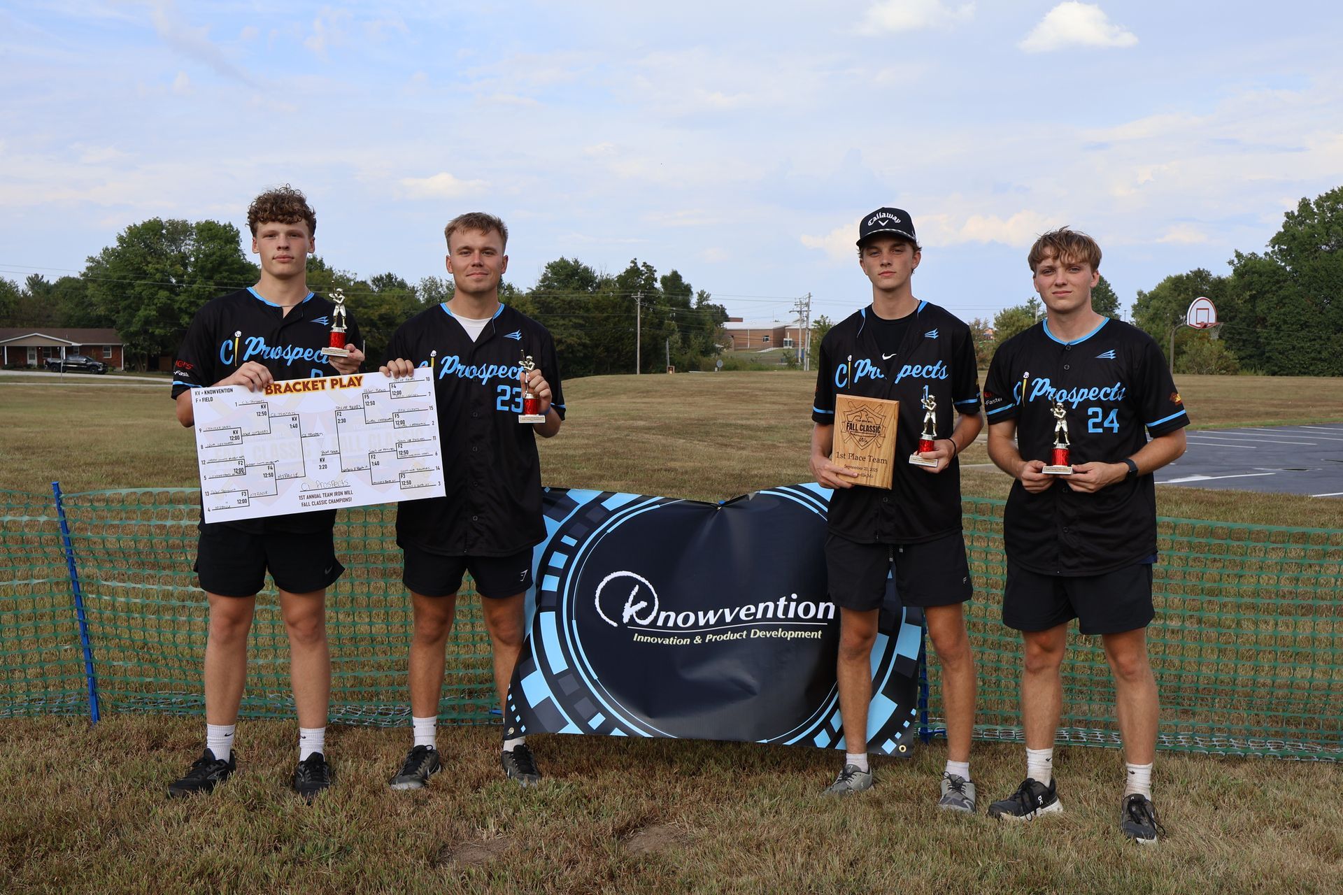 Group of four men holding trophies, standing on a wiffleball field. They are wearing black shirts and shorts, near a banner.