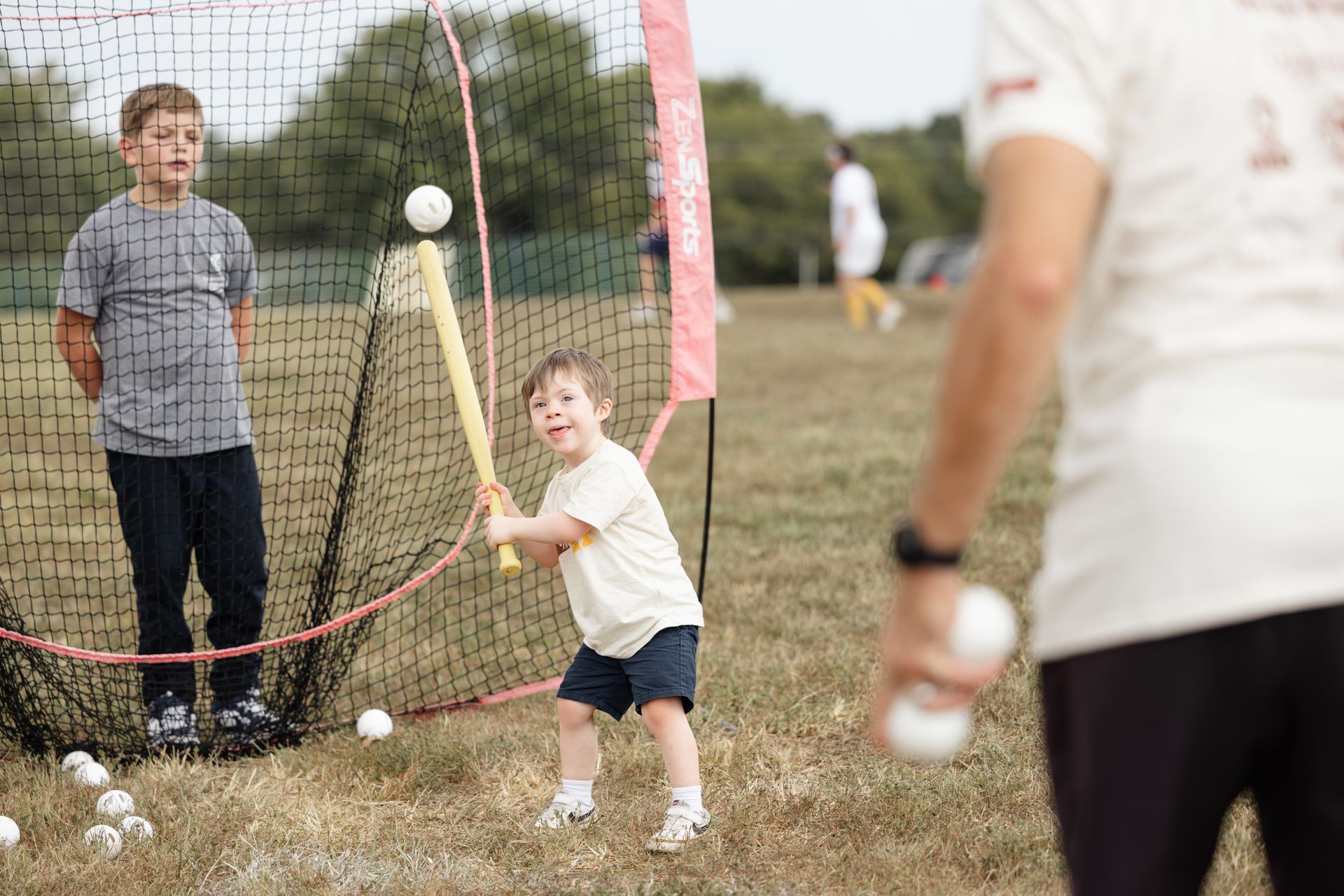 Boy swings a wiffleball bat, another boy watches, and a person throws balls outdoors near a net.