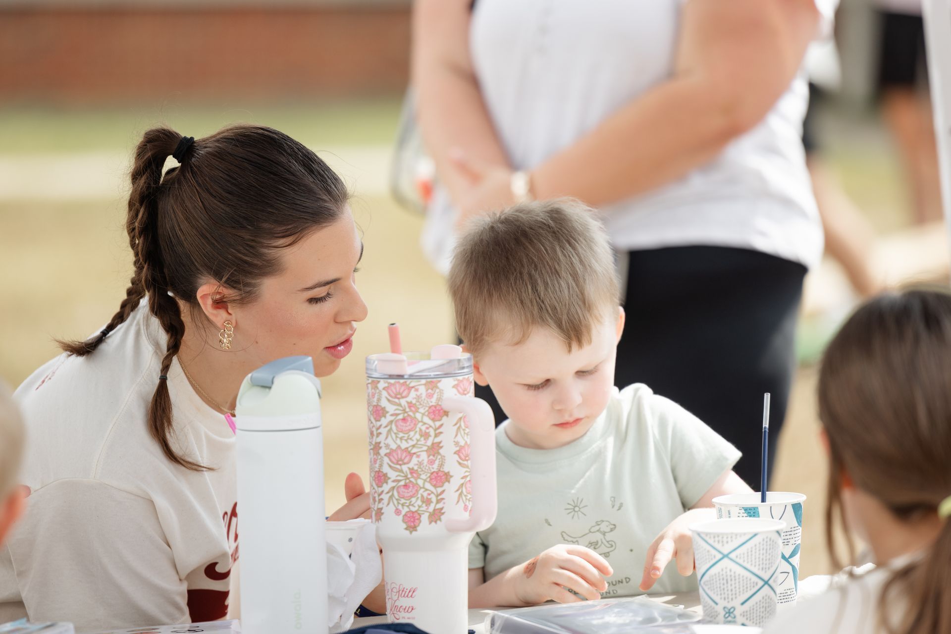 Woman helps a young child with a craft activity at an outdoor table.
