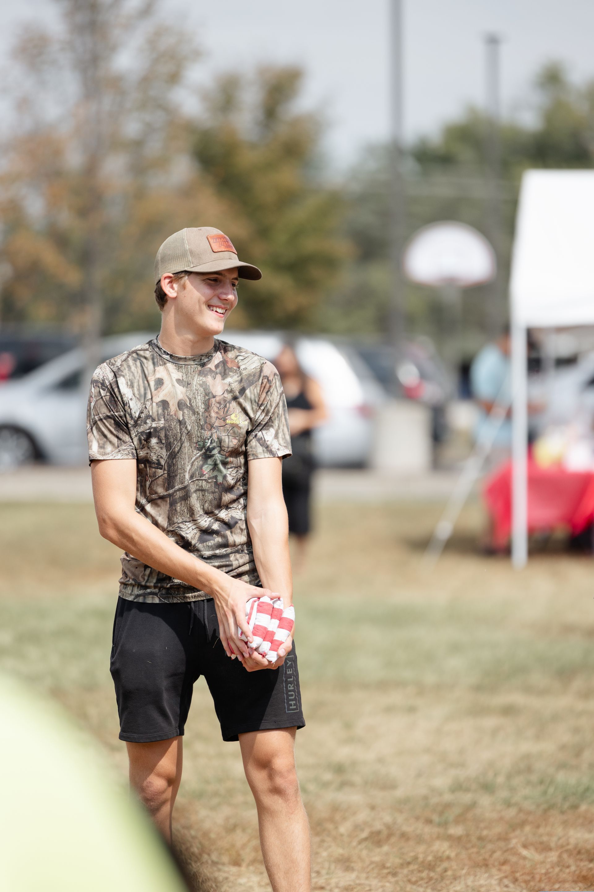 Man in camo shirt and hat holding ball, smiling outside on a sunny day.