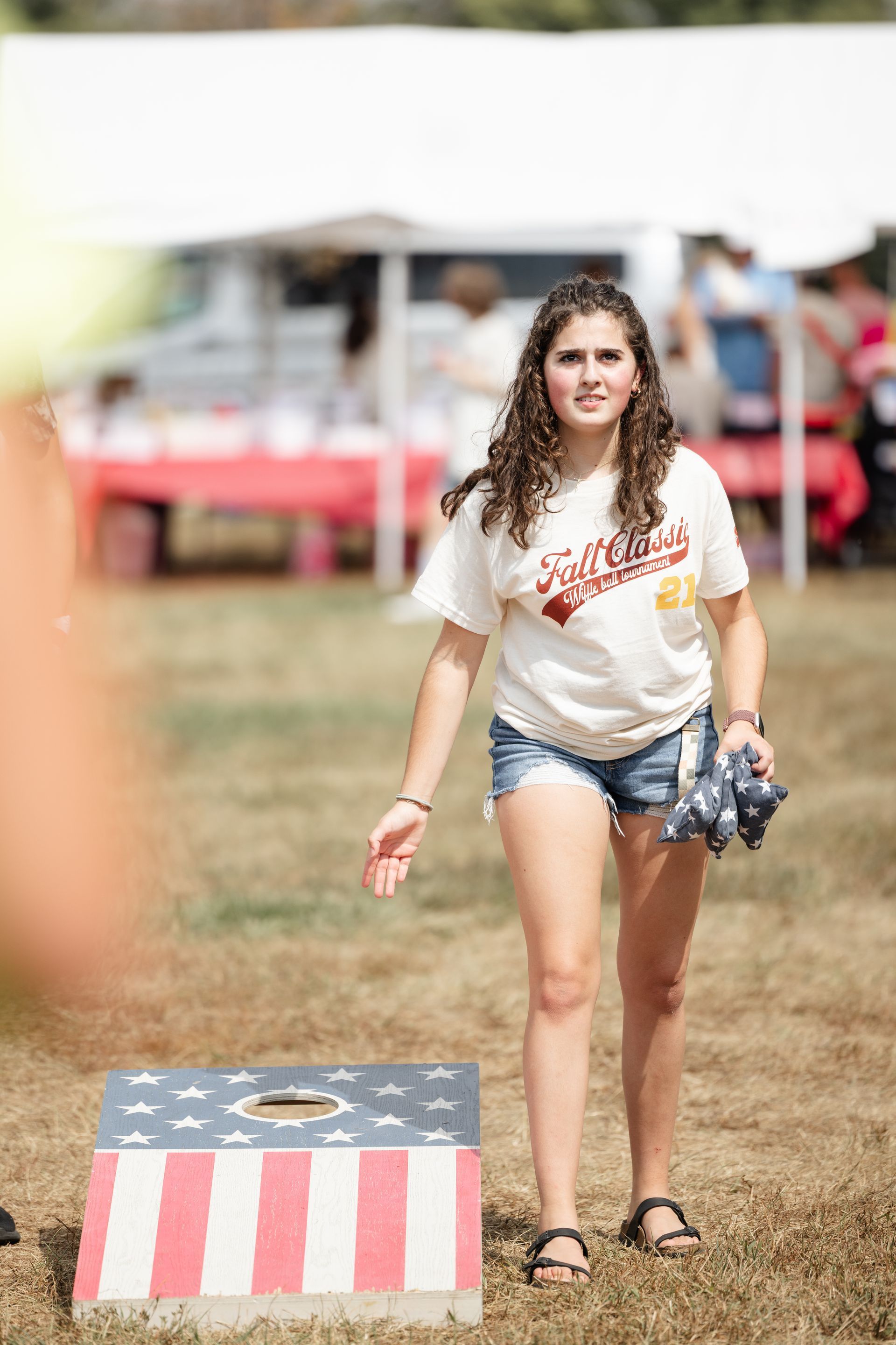 Woman in jean shorts and t-shirt throws beanbag at American flag-themed cornhole board on a grassy field.