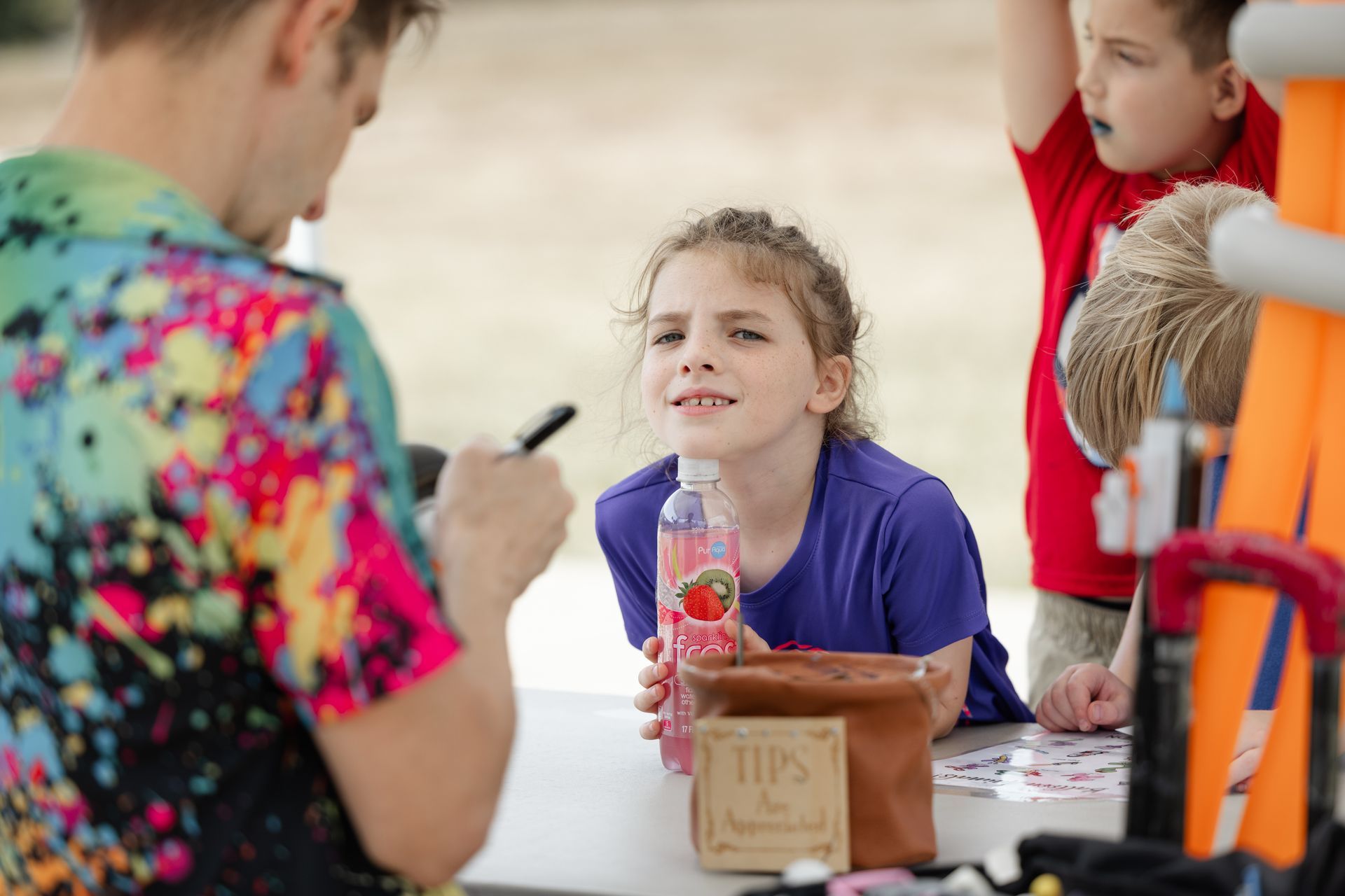 Girl at table with man writing; another child looks on. Outdoor setting, bright colors.
