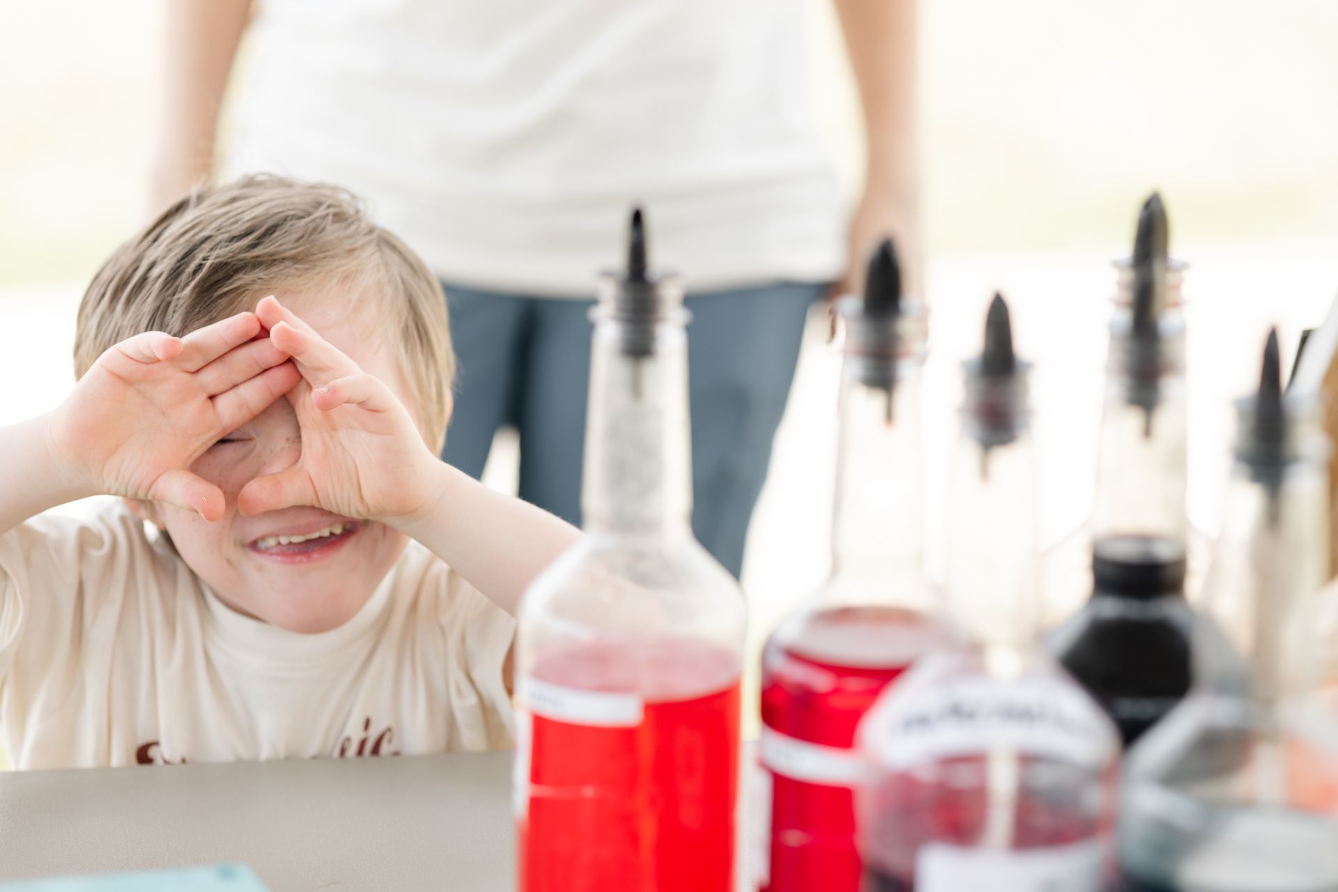Child (iron will) peeking through hands, smiling, in front of beverage bottles on a table.