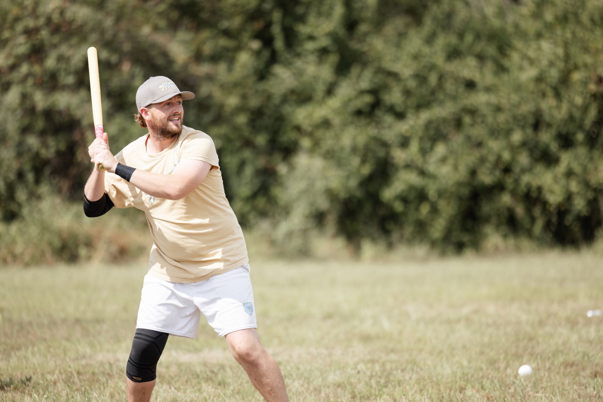 Man in a yellow shirt and white shorts swings a bat at a wiffleball in a grassy field.
